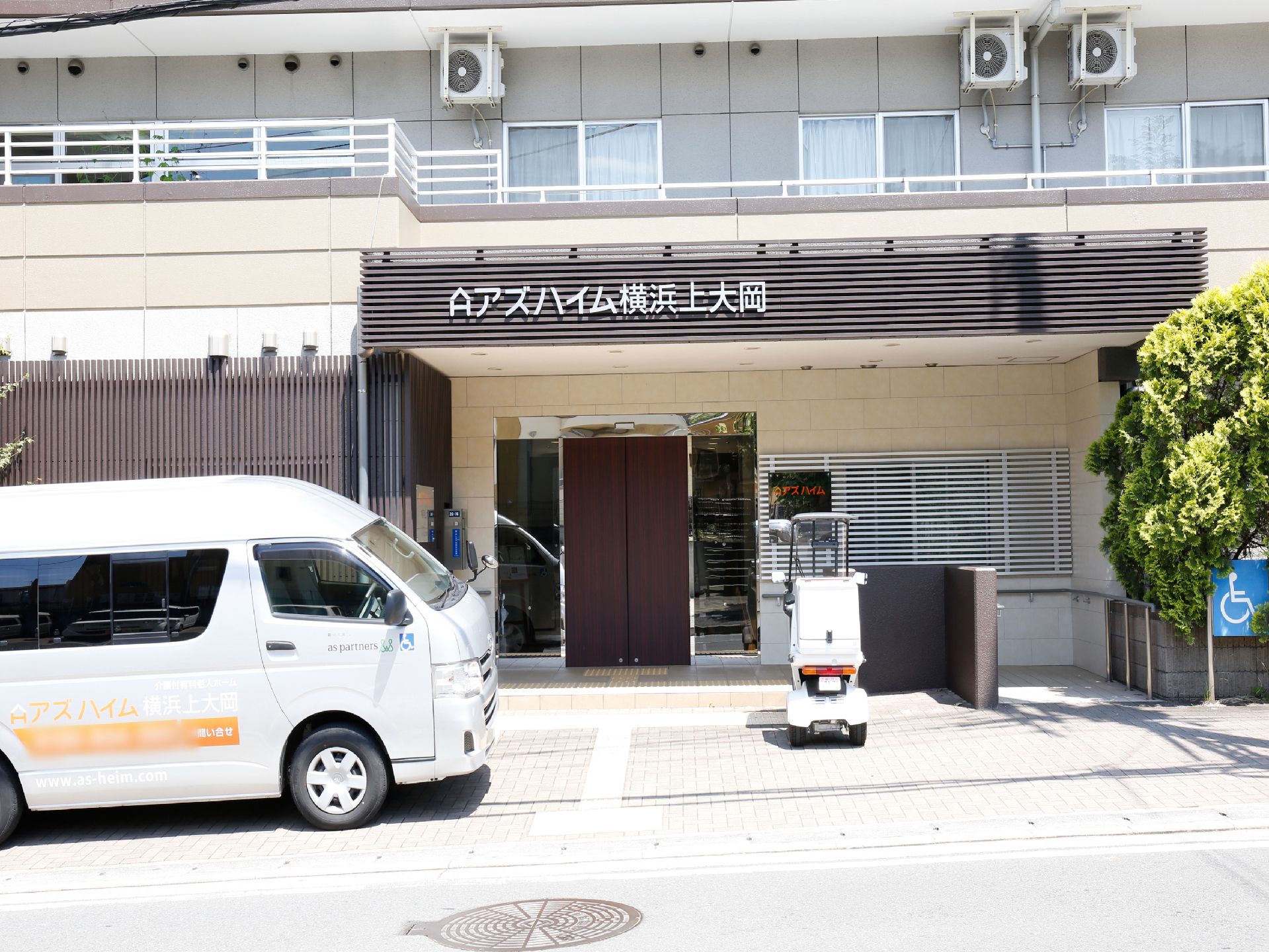 施設入口前には屋根がある。屋根の右側には手すり付きの通路がある。施設入口前には車とバイクが停まっている。