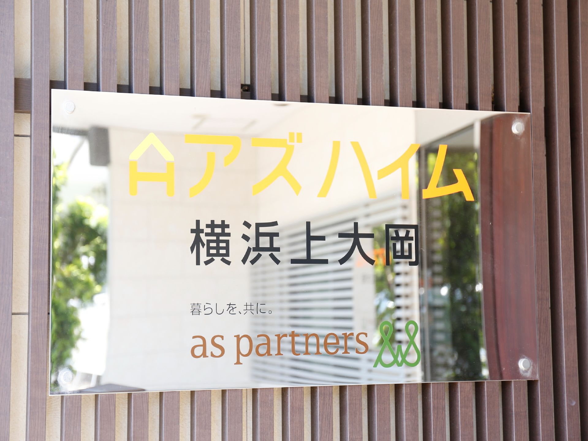 鏡のような素材の表札がある。表札には施設名と運営会社名が書かれている。施設名は黄色で書かれていて目立つようになっている。
