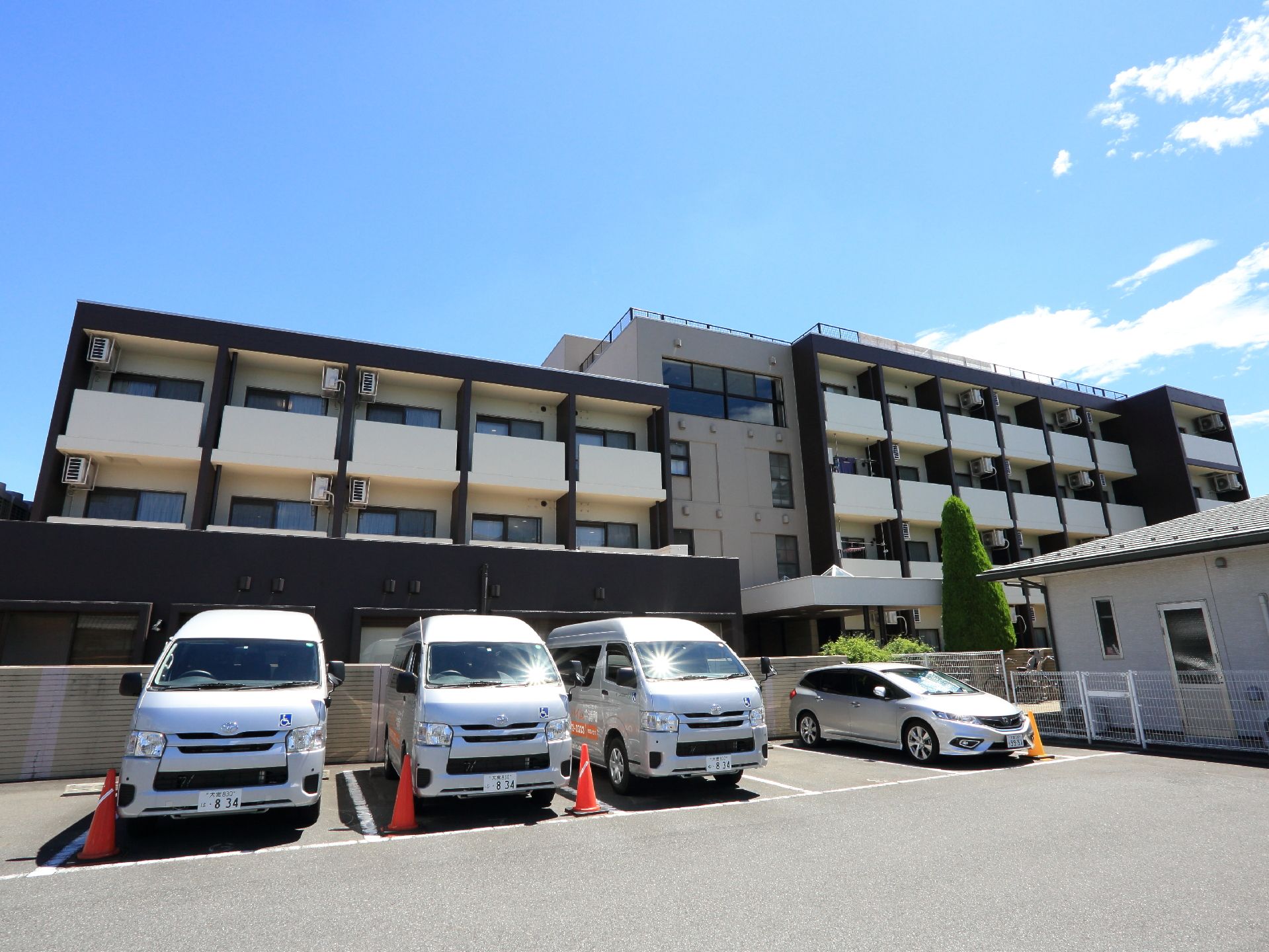 施設は落ち着いた色調の4階建て。敷地内には駐車場もあり、施設のワンボックスカー停められている。車いすマークが貼られている。