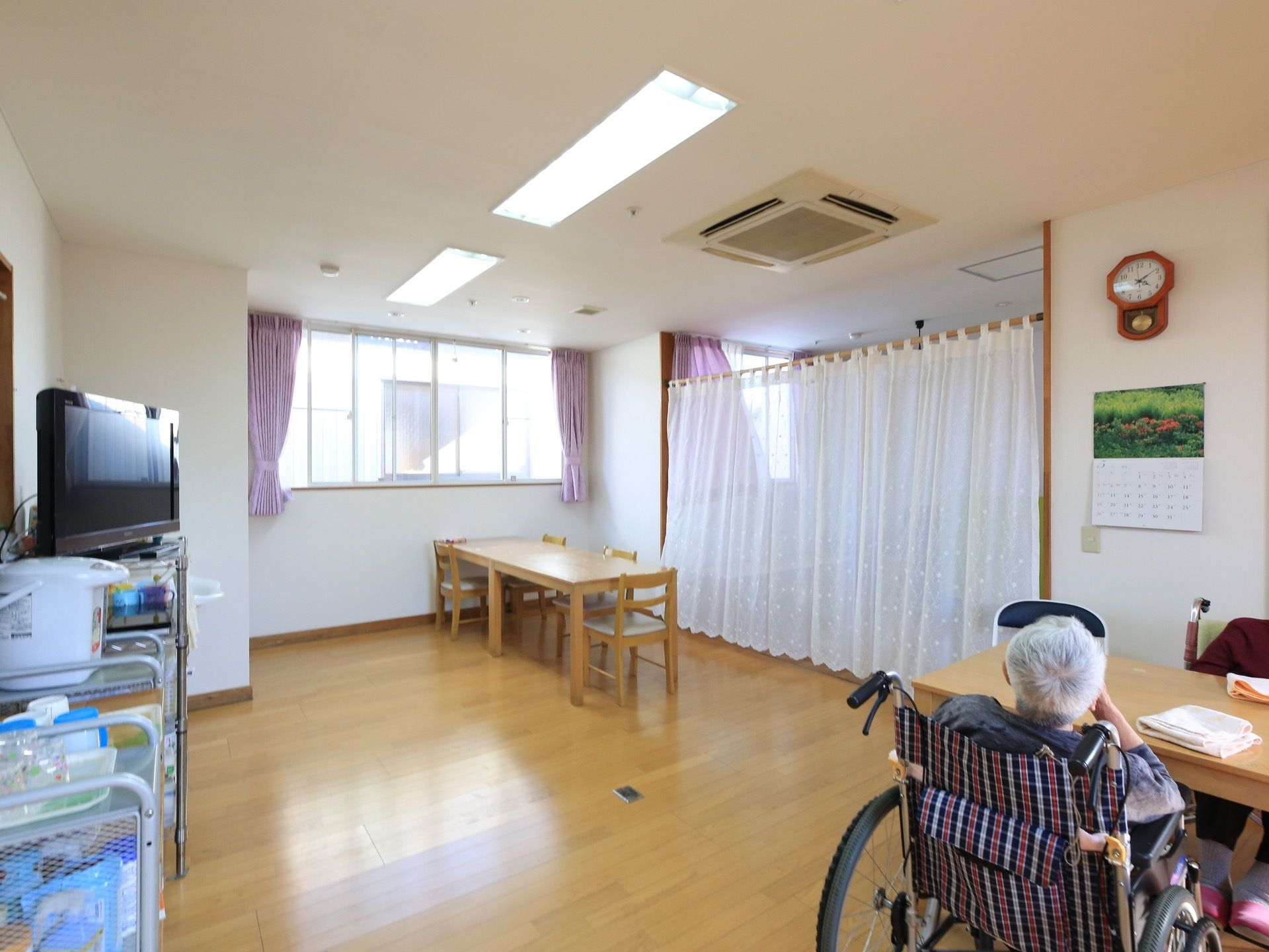 明るい木目調のフローリングの部屋に、テーブルや椅子、テレビなどが設置されている。奥の部屋とはレースカーテンで仕切られている。