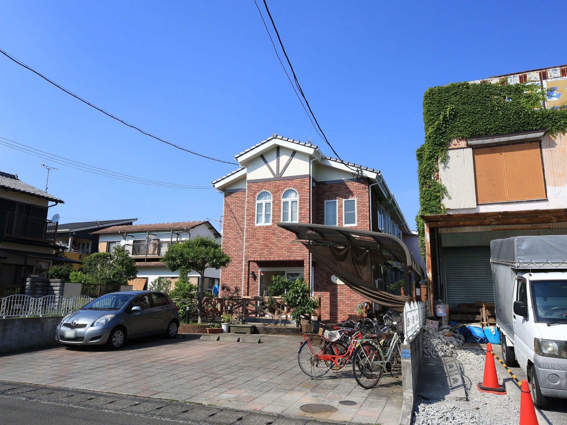 三角屋根が印象的なレンガ調造りの施設建物の様子である。前面には植栽が施され、駐車場や自転車置き場が設けられている。