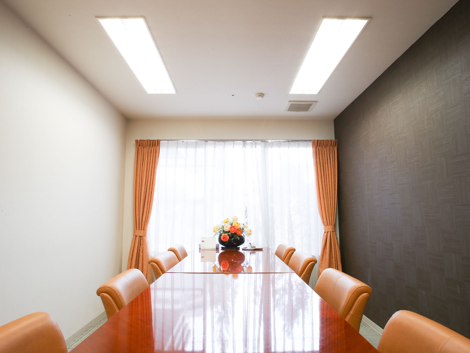 部屋にはカーテン付の窓がある。茶色いテーブルが2台あり、周りには椅子が8脚置かれている。テーブルの上には花瓶がある。