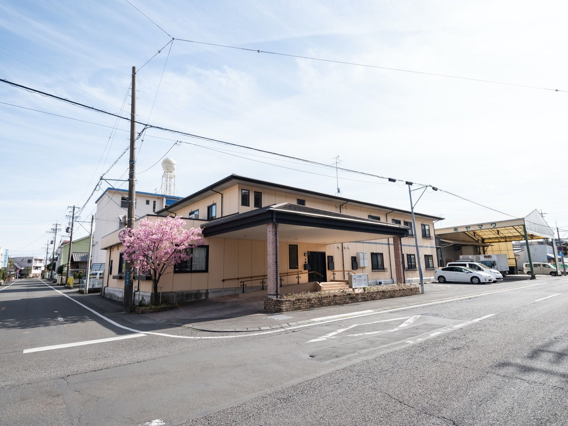 家庭的な雰囲気の施設外観である。2階建てになっており、敷地内には桜の木が植栽されている。駐車場には車が2台停められている。