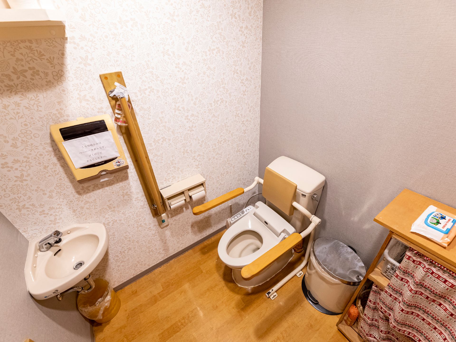 原っぱのような場所でブルーのエプロンを付けた女性スタッフに付き添われて歩いている高齢女性。女性はブラウンのジャケットを着ている。