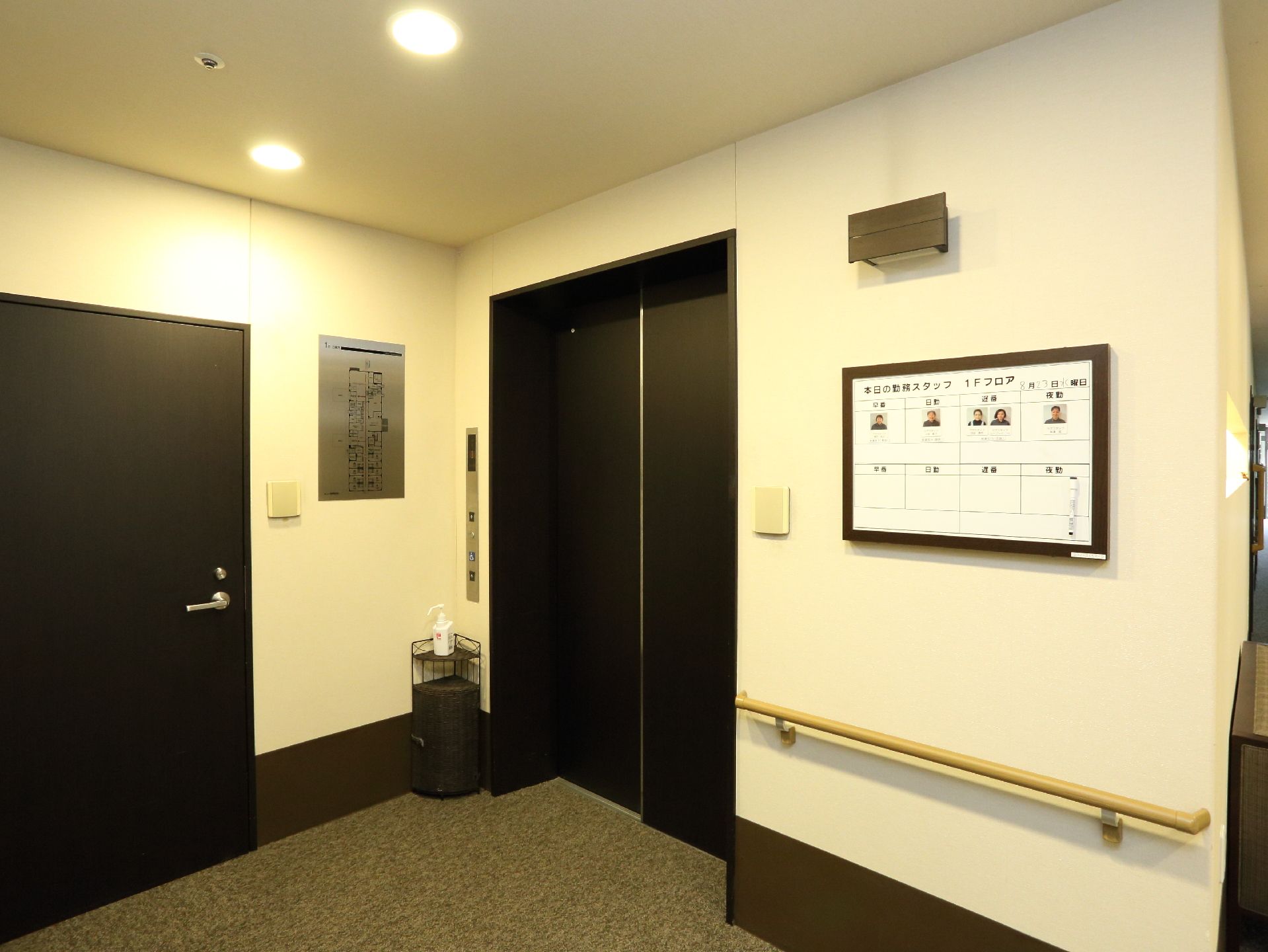 エレベーターの横に、今日施設にいるスタッフの名前と写真が掲示されている。壁には手すりが設置されている。