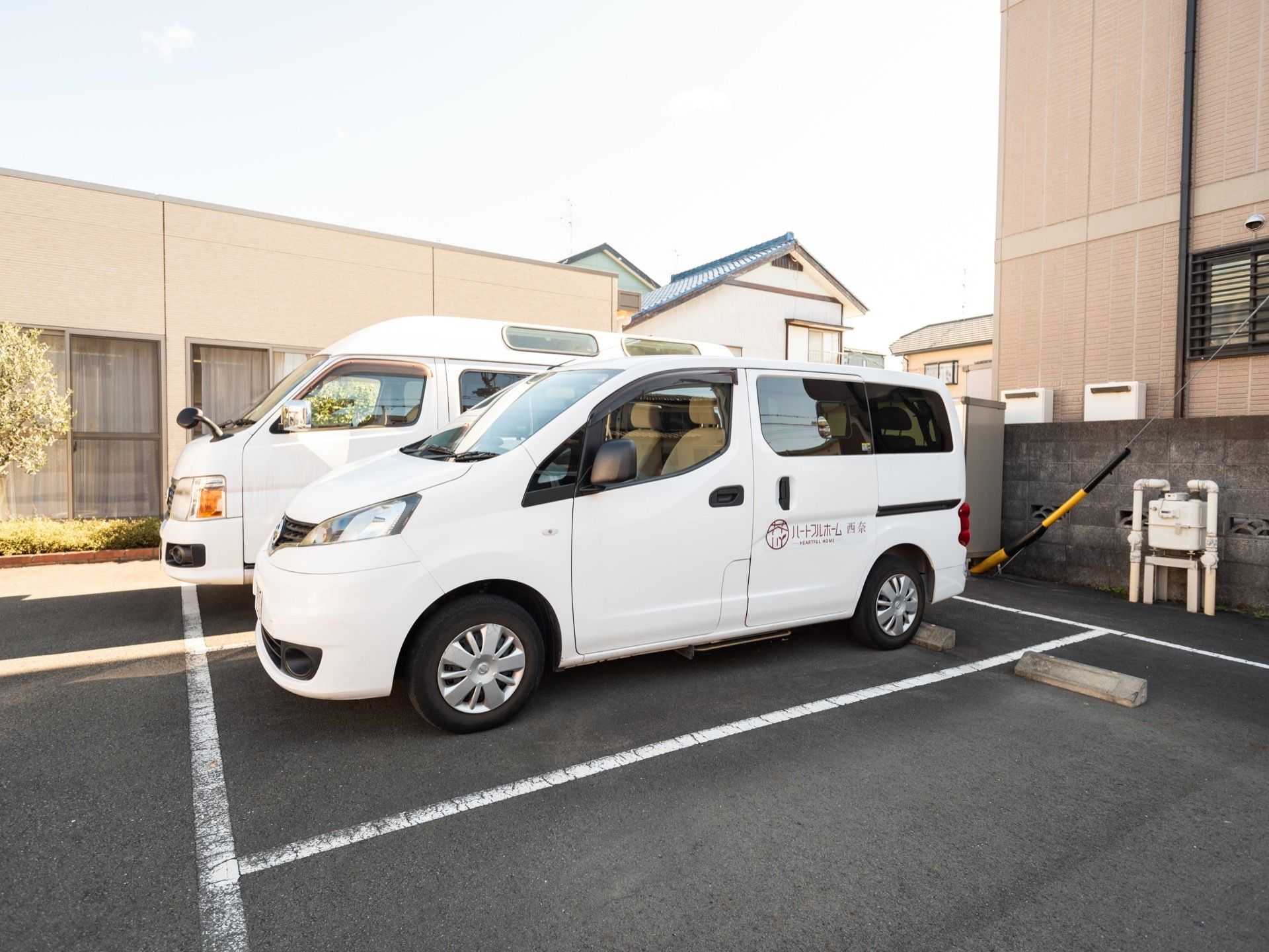 淡いホワイトカラーに施された施設外観である。植栽が施されている。施設名が記された福祉車両が2台停められている。