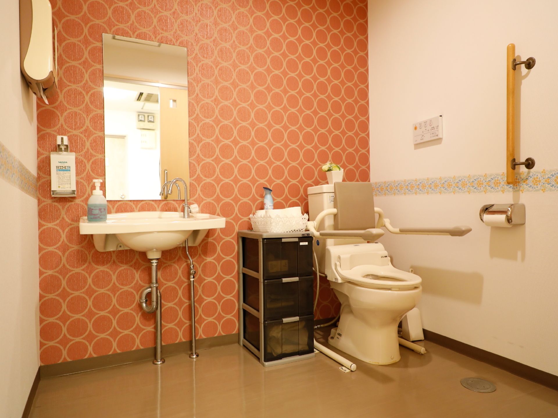 温水洗浄暖房便座トイレで、操作パネルは壁に取り付けてある。洗面台、手指用消毒スプレー、鏡、収納ケース、ペーパータオルがある。