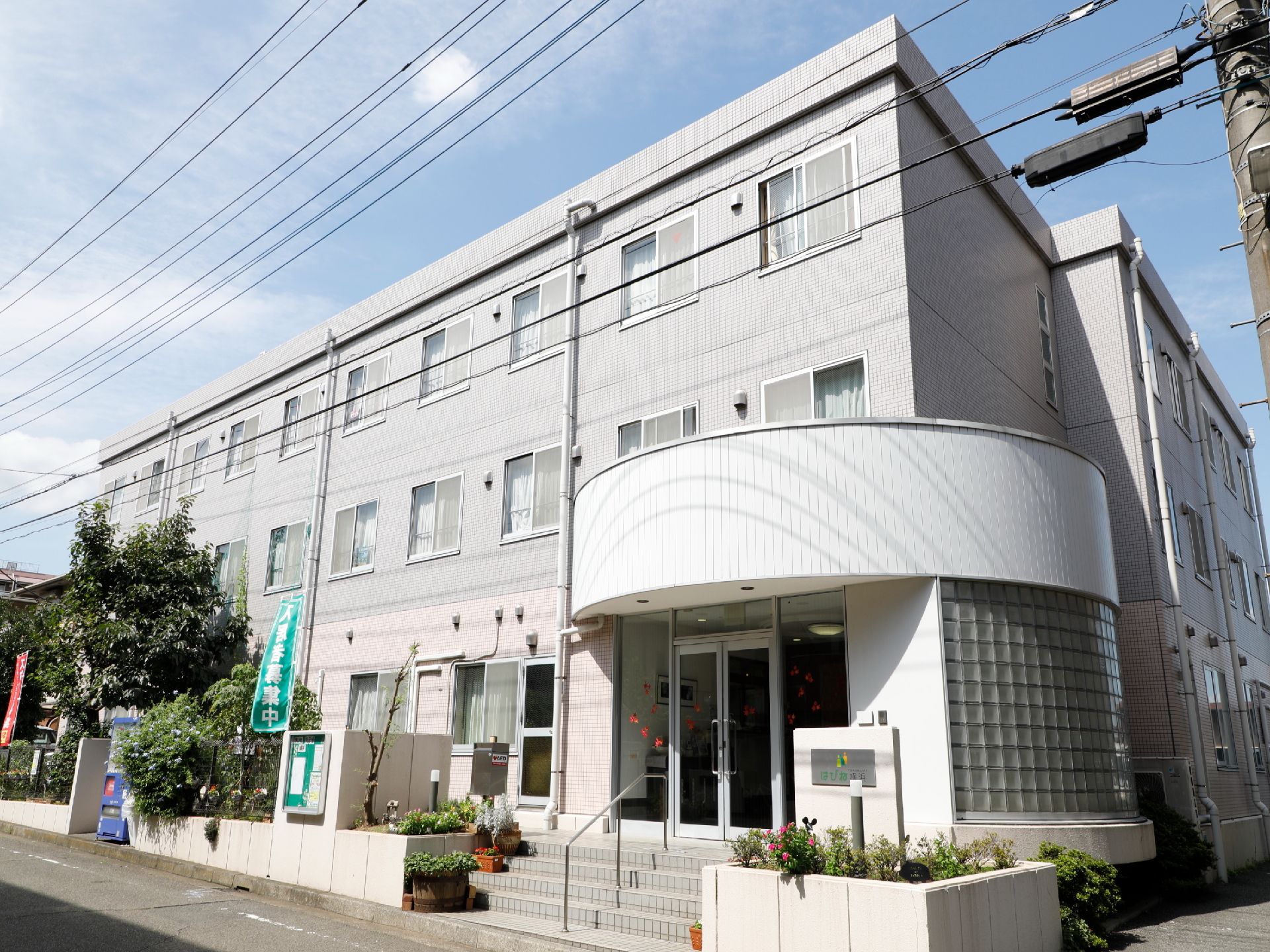施設は3階建てである。玄関前の階段には手すりが備わっている。入居者様募集中と書かれた緑色ののぼりが立てられている。