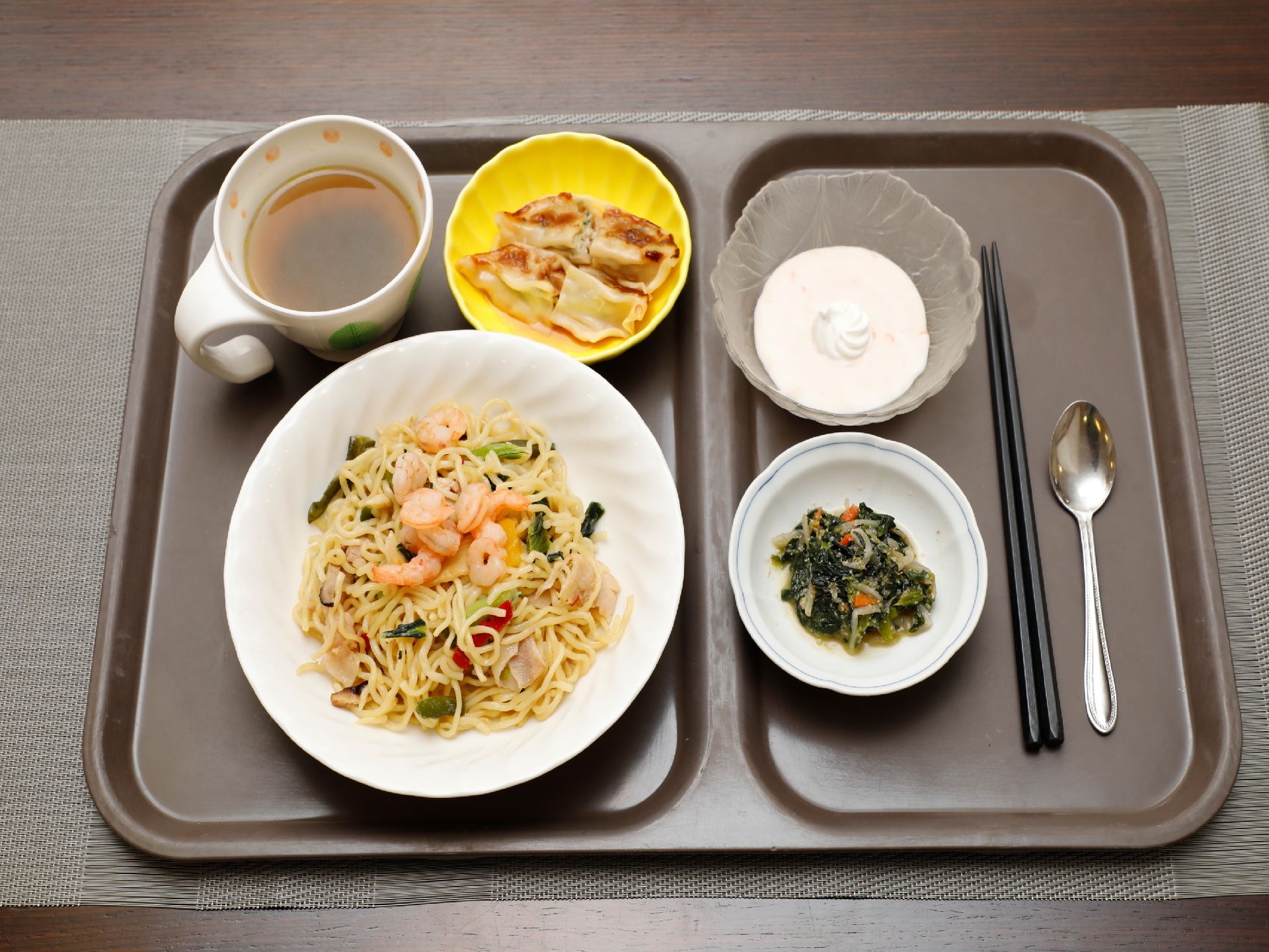 この日の食事が食器に盛りつけられ、茶色のトレーに乗せられている。焼きそば、小鉢2品、デザートが用意されている。