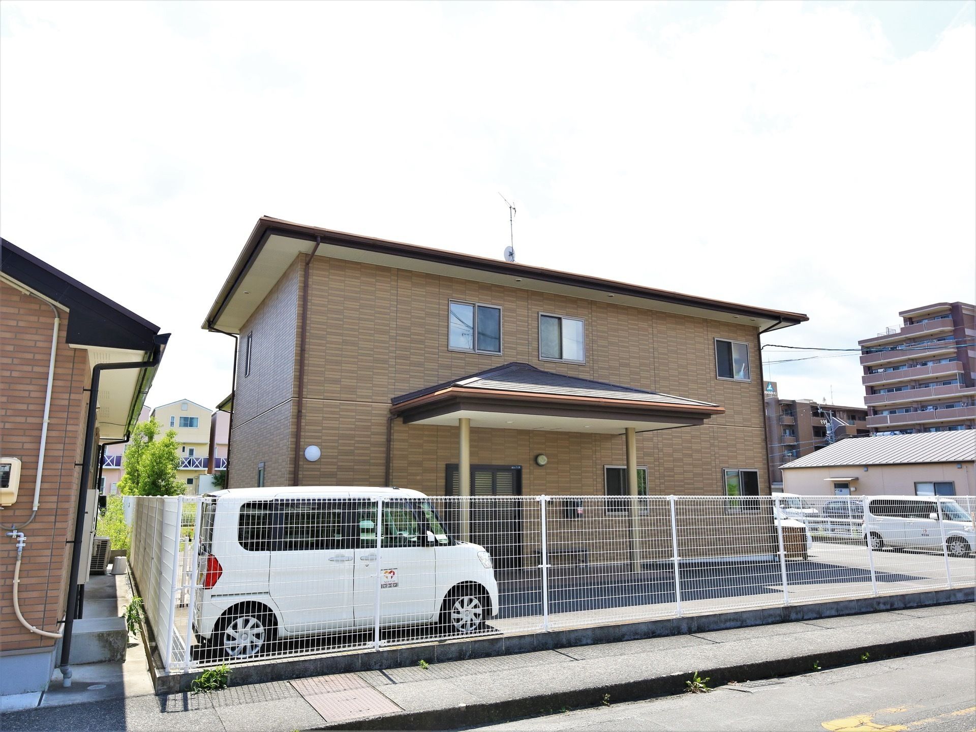 茶色を基調としたタイル調の壁が特徴的な建物である。道路と敷地は白いフェンスで区切られ、敷地内に車が駐車されている。