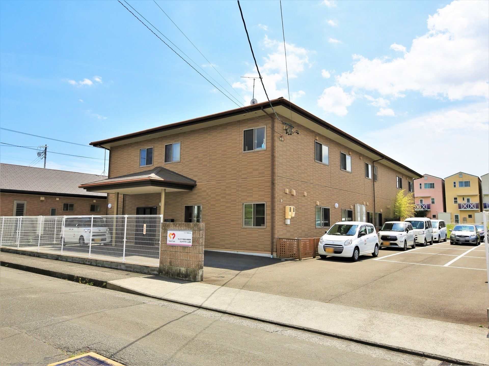 施設建物は鉄骨２階建てで、茶色を基調とした壁が特徴的である。敷地内には施設が所有する車が複数停められている。