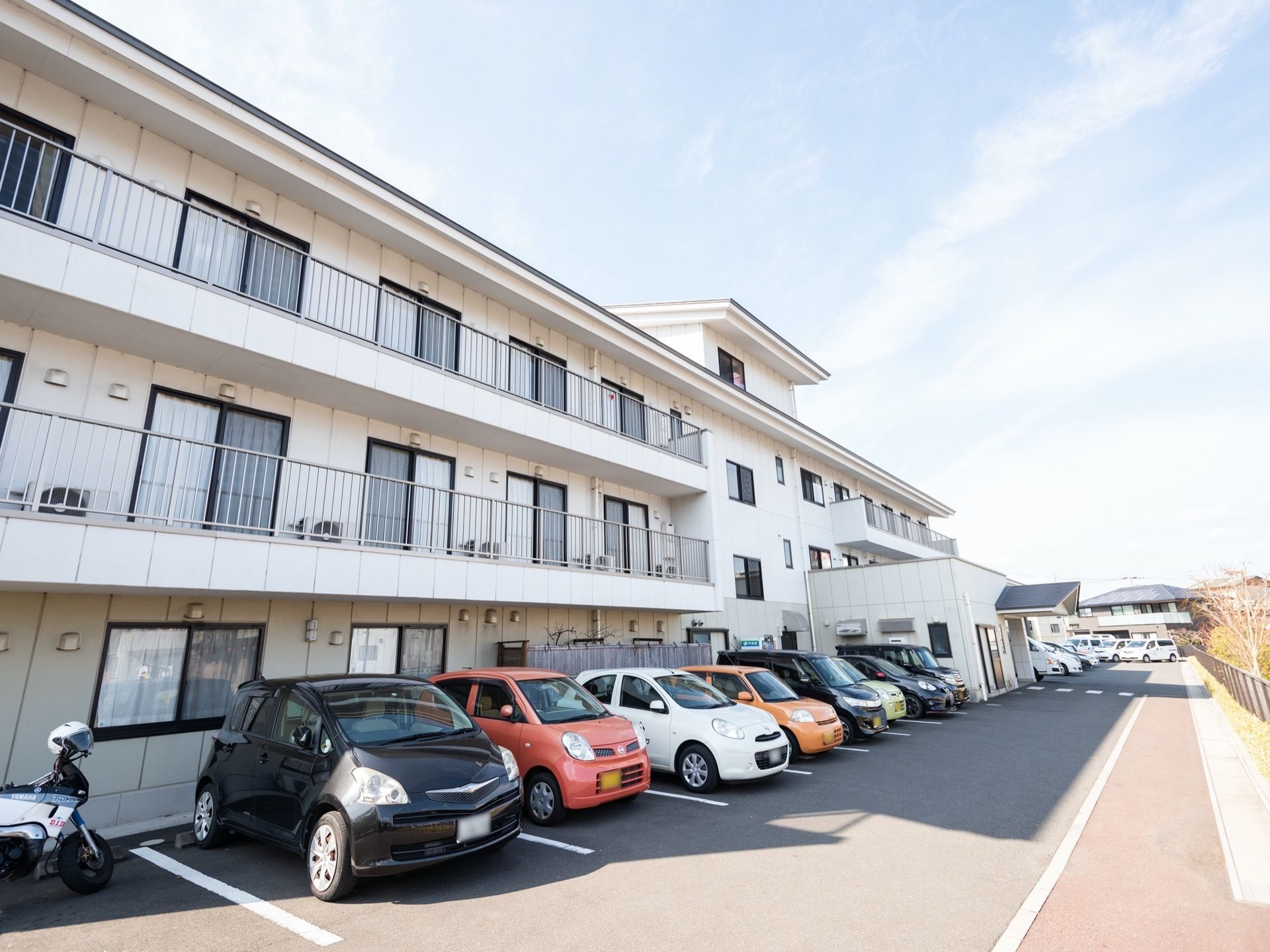 3階建てのアパート風の建物である。前個室にバルコニーが設置されており、駐車場には車が停められている。