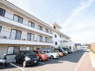3階建てのアパート風の建物である。前個室にバルコニーが設置されており、駐車場には車が停められている。