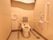 柔らかい照明器具の個室のトイレがある。便座にはひじ掛けと背もたれが付いており、壁には縦方面に手すりがある。