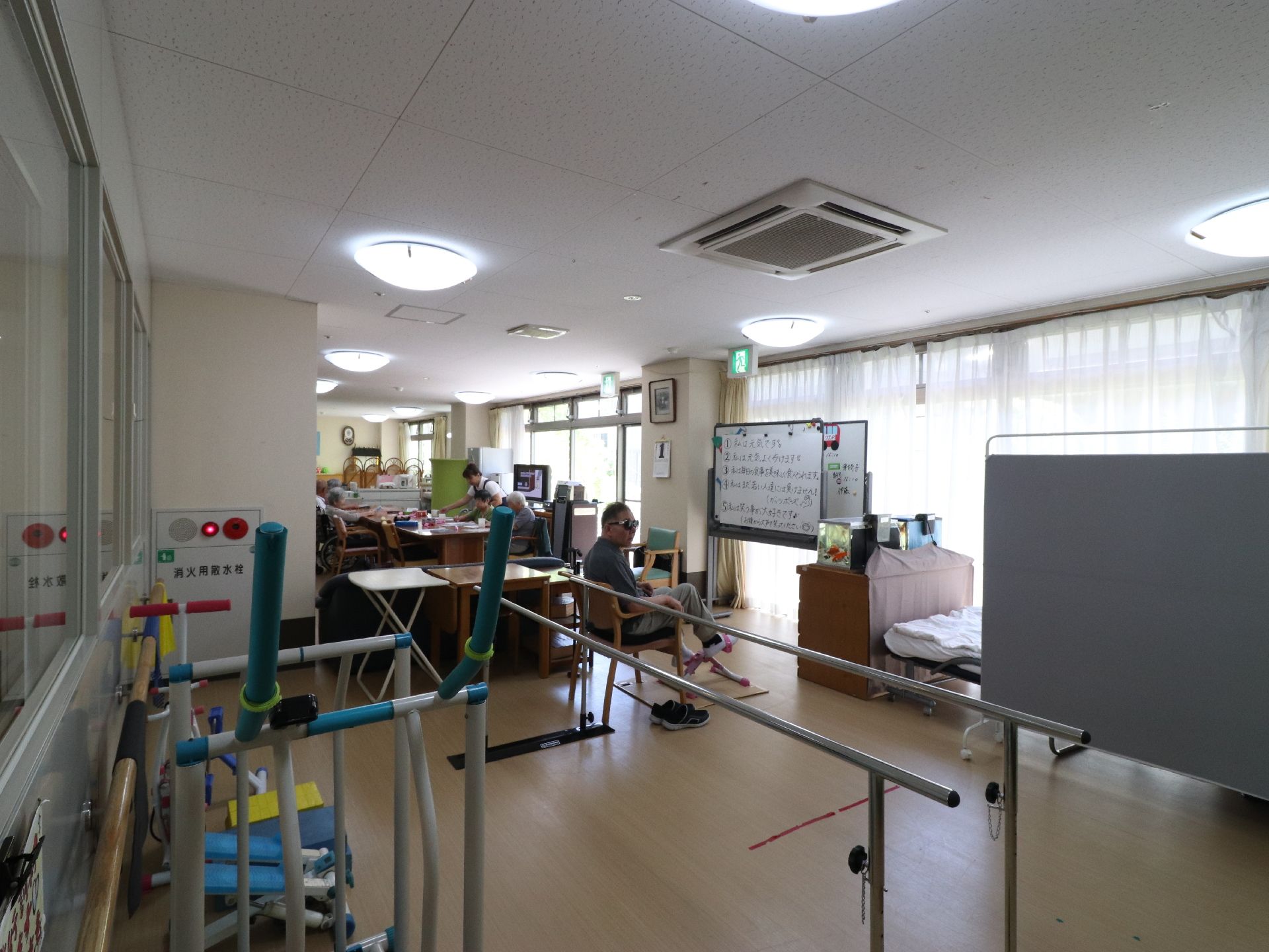 歩行訓練用のバーの他、運動用の機材が置かれている。窓側にはベッドがある。奥には机と椅子、テレビなどがある。