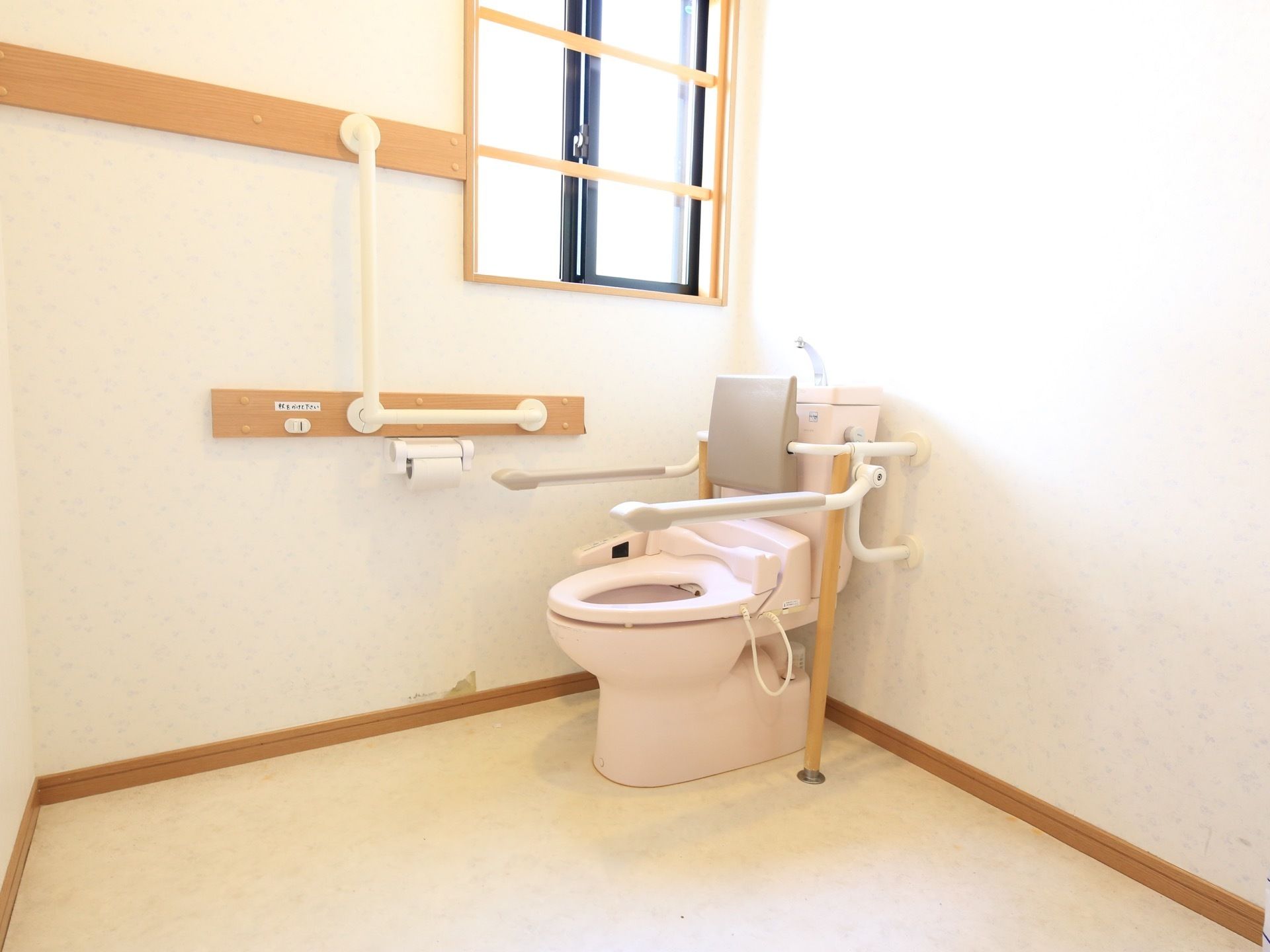 白い壁に囲まれた広いトイレがある。大きな窓が付いており、木製の柵がはまっている。壁には手すりがあり、便座にはひじ掛けが付いている。