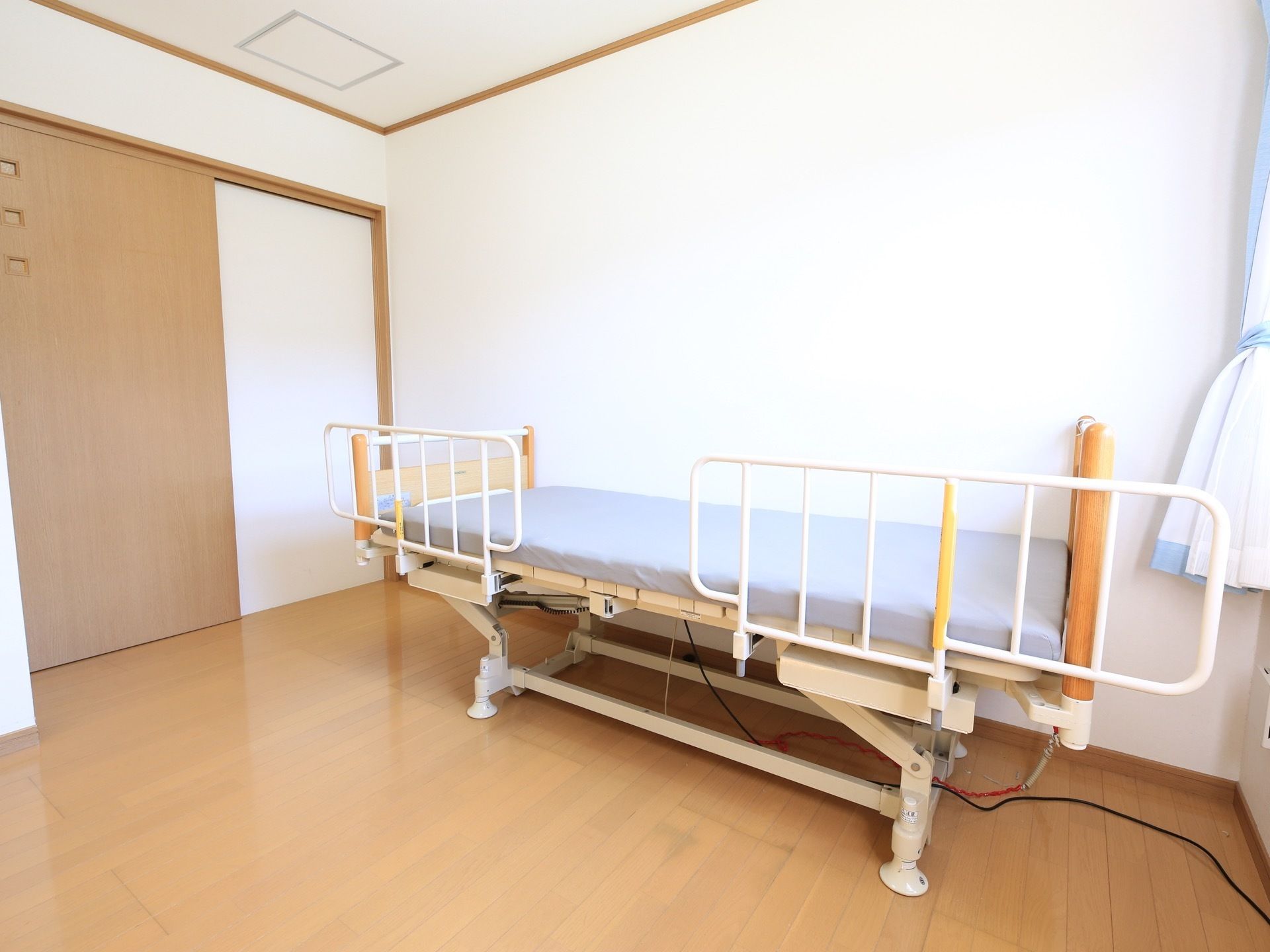 ライトブラウンの床と白い壁の居室がある。壁際には柵の付いた介護用ベッドが置かれ、水色のマットレスが敷かれている。