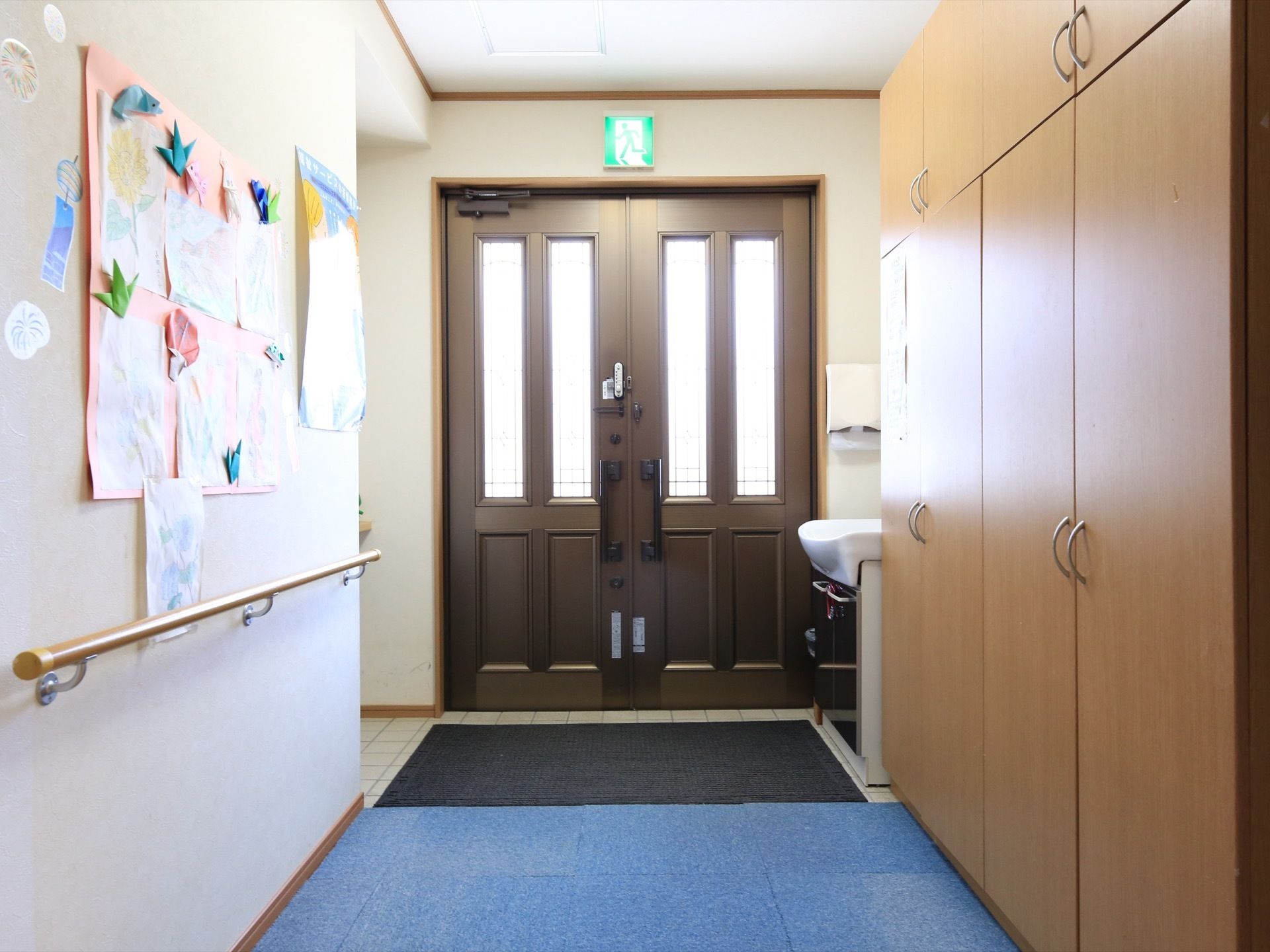 ダークブラウンの両開きの玄関ドアがある。すぐ側には洗面台と備え付けの大きな棚がある。足元には玄関マットがある。