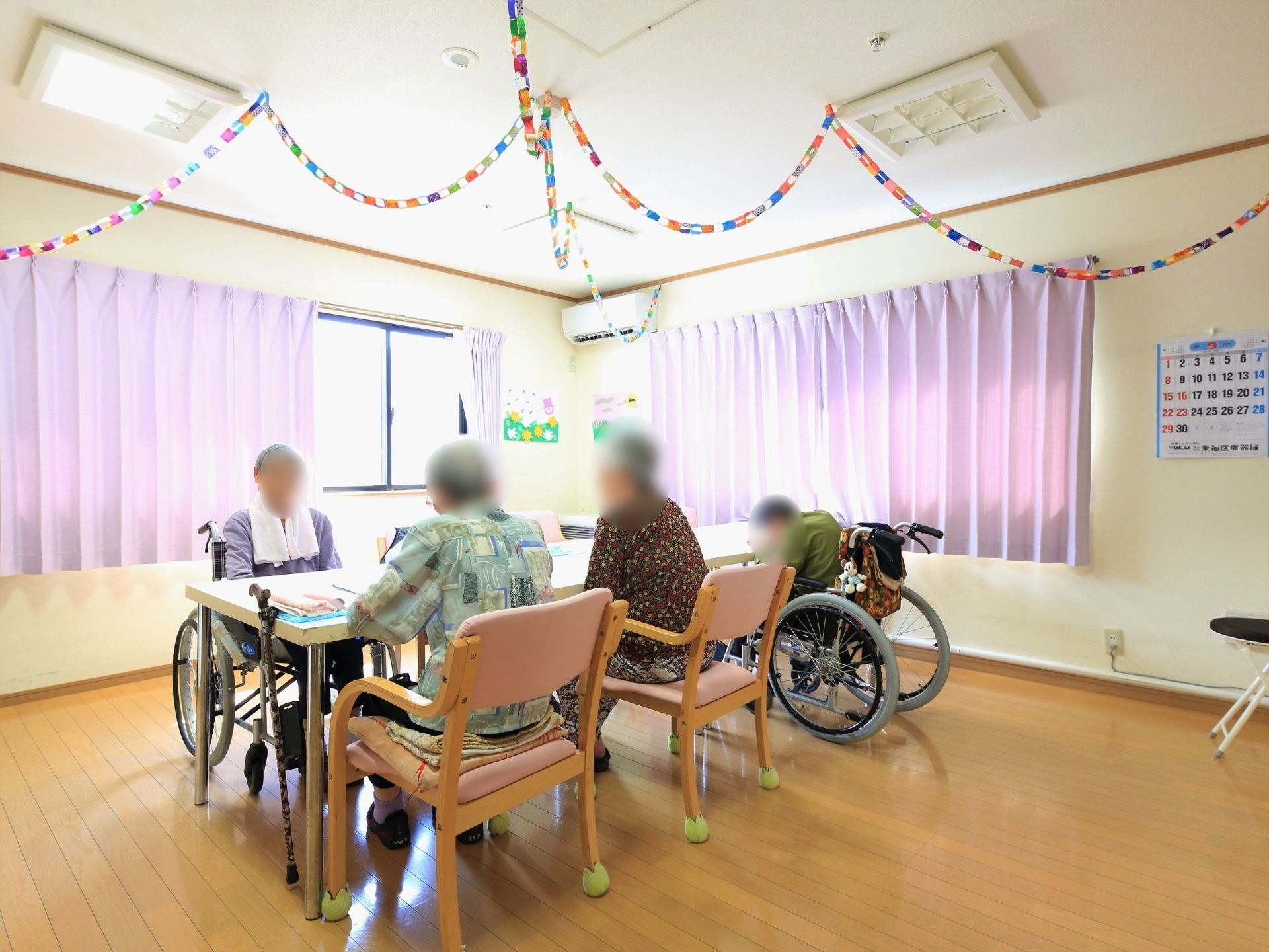 ライトブラウンの床の明るい雰囲気の談話室。中央に大きなテーブルがあり、入居者様が座られている。天井には手作りの飾りつけがされている。