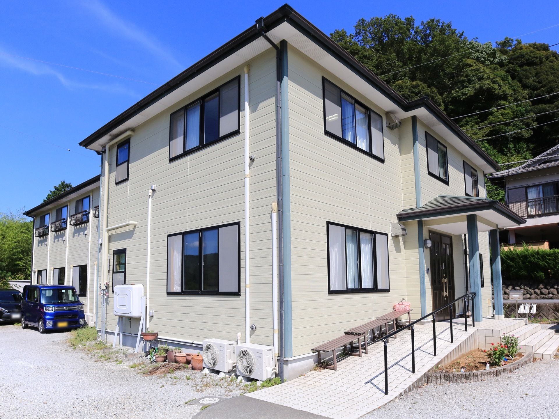 ライトグレーの壁の、２階建ての建物がある。玄関に向かってスロープと手すりがある。建物の背後には大きな木々が繁っている。
