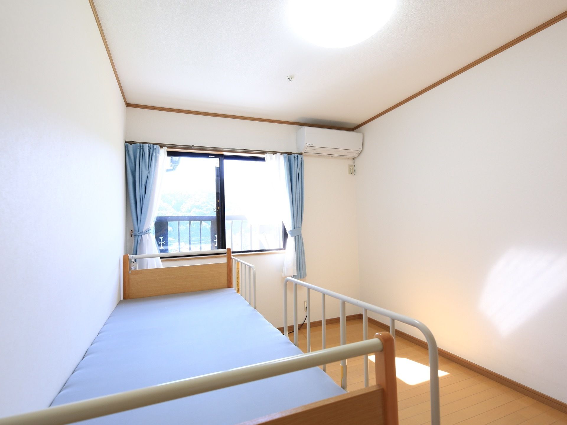 明るい色調のフローリングと白い壁の居室がある。カーテンの掛かった窓があり、介護用ベッドがある。