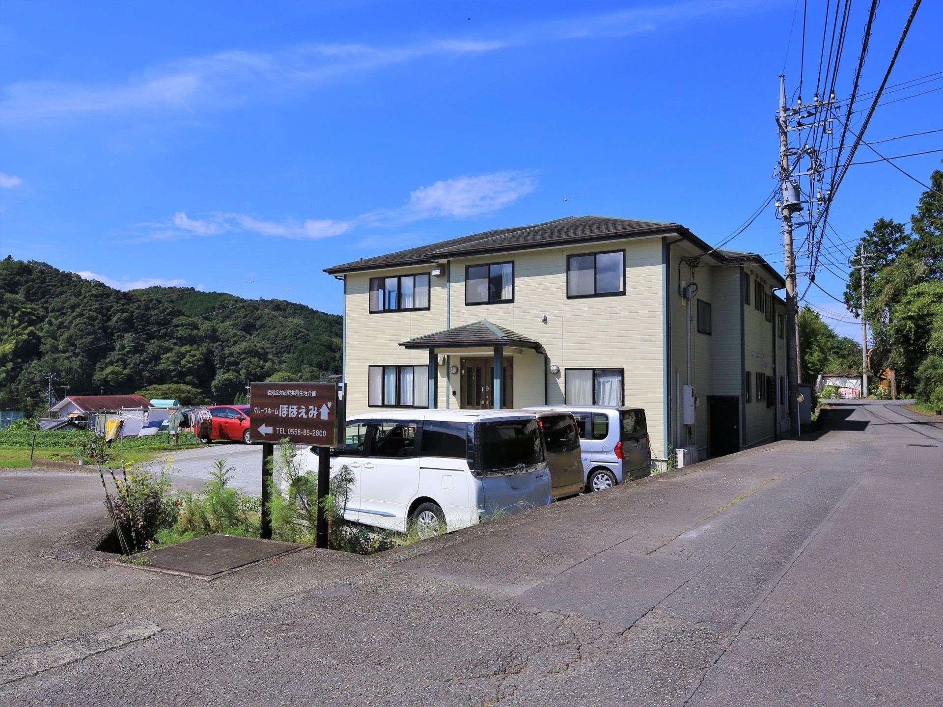 道路沿いに建っている２階建ての建物がある。周囲は緑に囲まれ、自然豊かな環境である。敷地内には駐車場があり、車が数台停まっている。