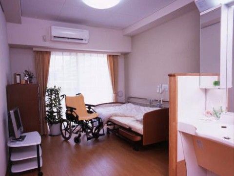 白と木目調の落ち着いた雰囲気の居室。エアコンとテレビ、介護ベッドもあり、手前には鏡付きの洗面台も設置されている。