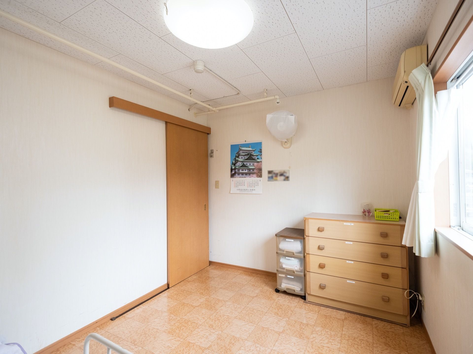 日当りがよく明るい居室である。暑い時期に備えてエアコンのほかに扇風機も設置されている。使用しない時にはカバーを掛けている。