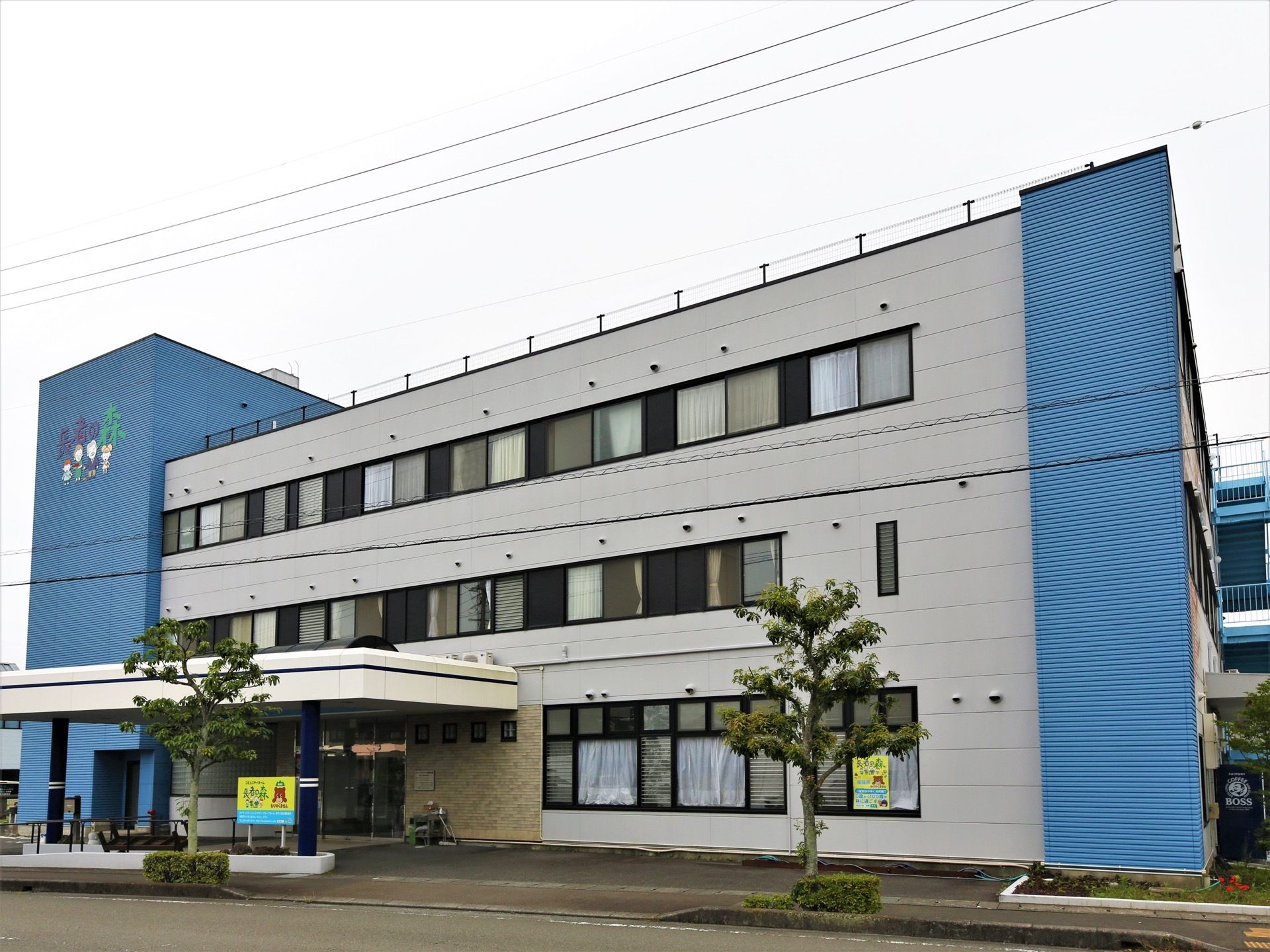 明るめのグレーとブルーで彩られた、３階建ての施設である。目の前は道路が走っている。複数の電線も見える。