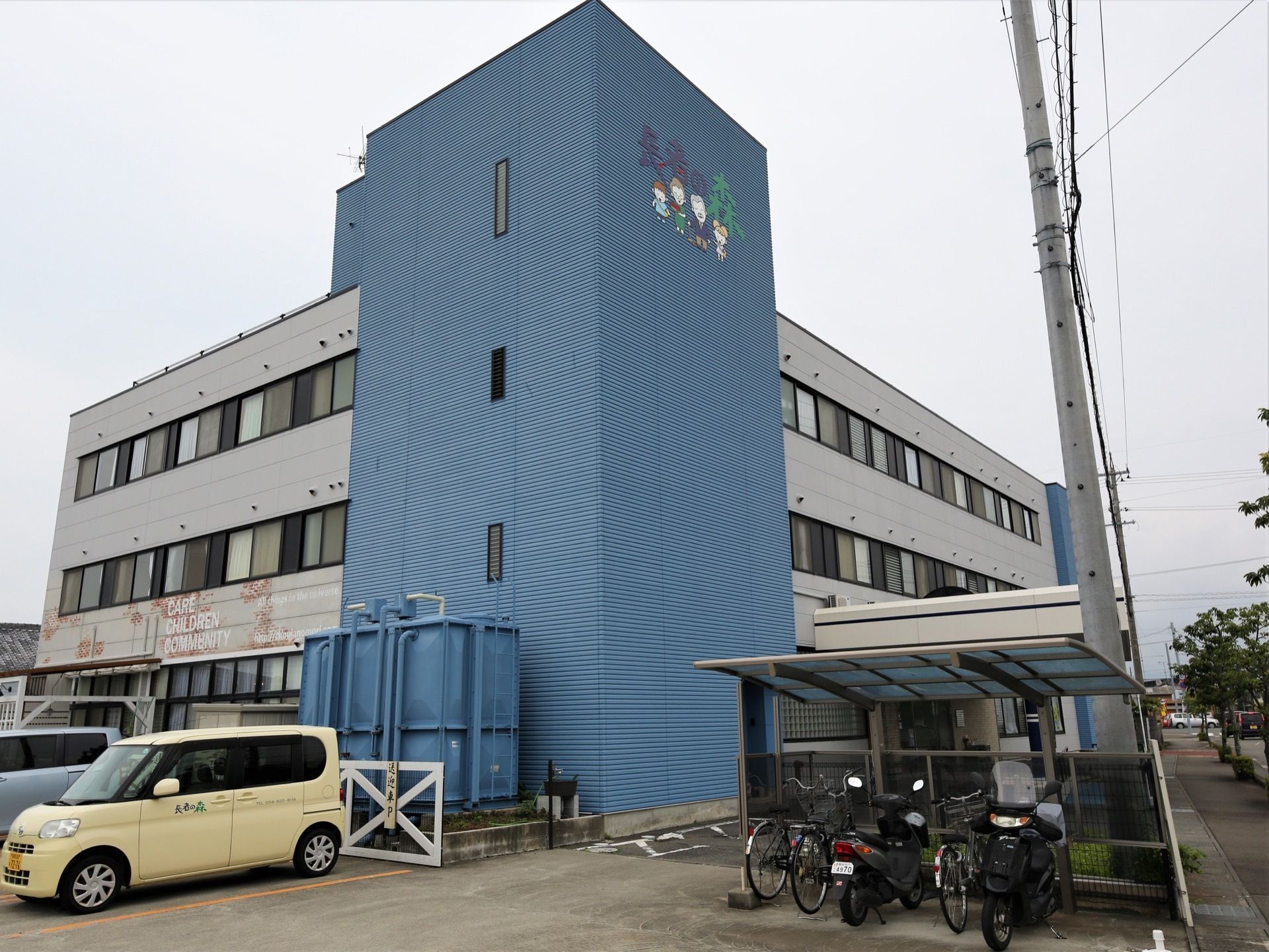 ブルーの外壁が印象的な建物である。電柱のそばに駐輪場が設置されている。ラインが引かれた駐車場もある。