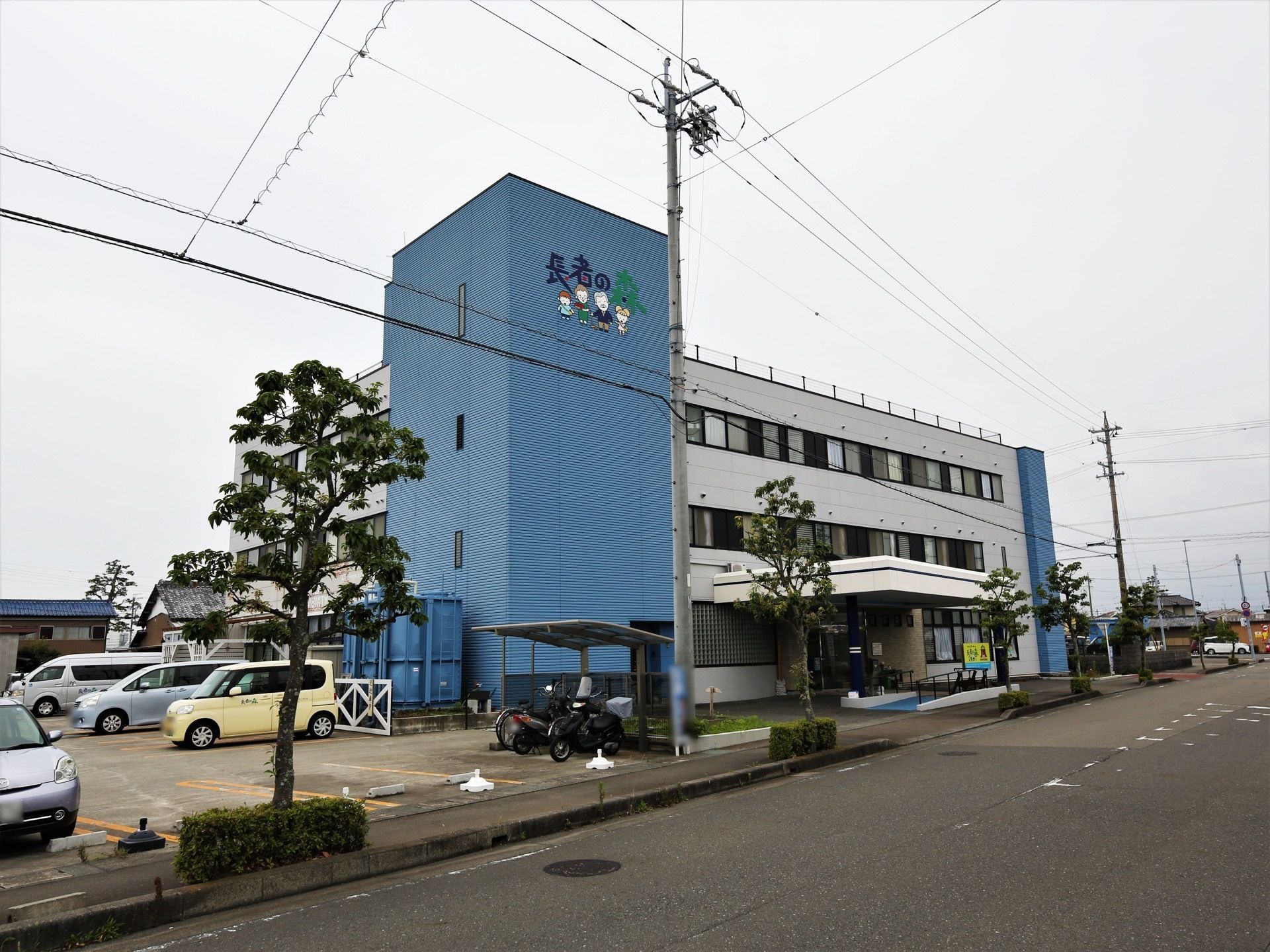 道路に面した場所に立地する、存在感のある施設である。歩道の随所には木々が植えられている。電柱も立ってある。