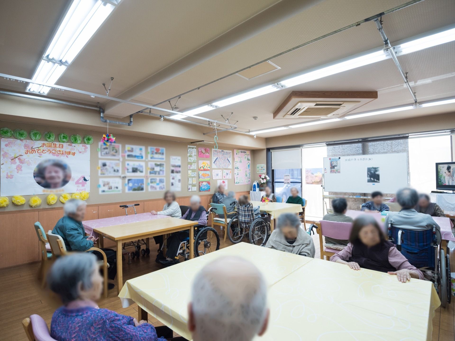 広く造られた共有スペース。壁面には写真やお知らせなどの掲示物が貼られている。２台合わせたテーブルがあり、椅子や車椅子に入居者様が座っている。