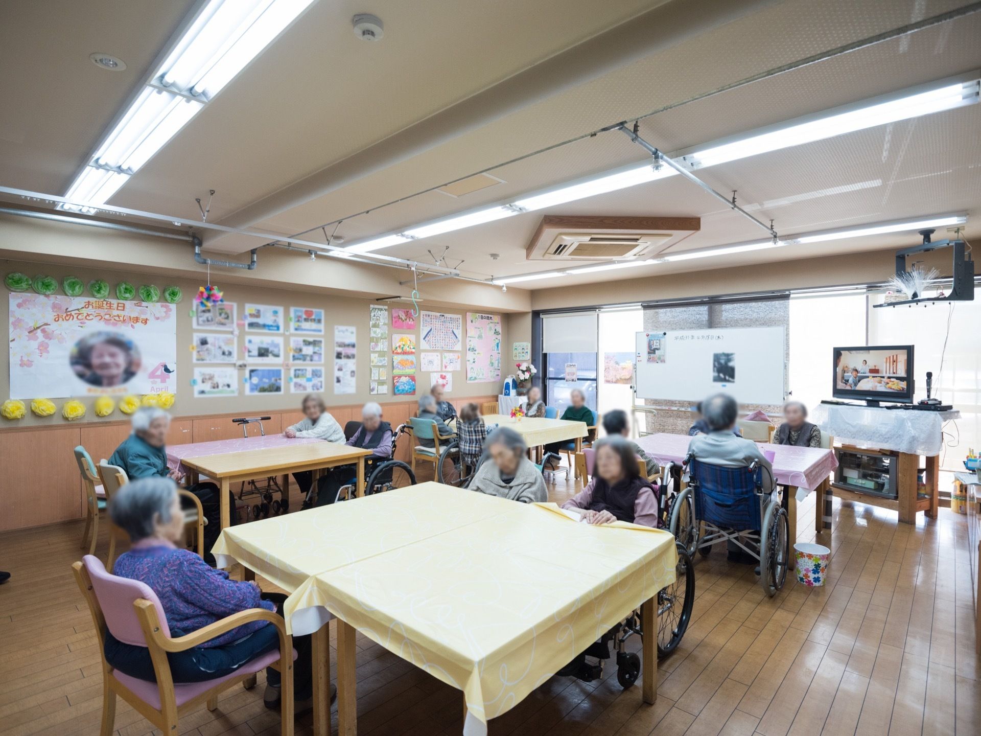 テーブルを囲むように向かい合って座っている入居者様の姿が見える。テーブルには、ビニール製のクロスが掛けられている。