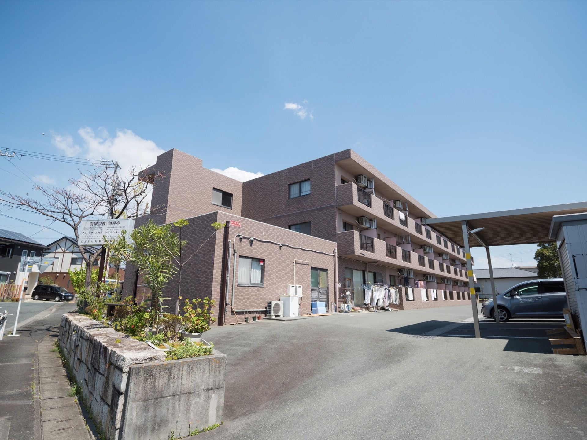 横幅の広い３階建ての重厚な施設の前面に、アスファルトの駐車場が広がっている。周辺には住宅が建ち並んでいる。