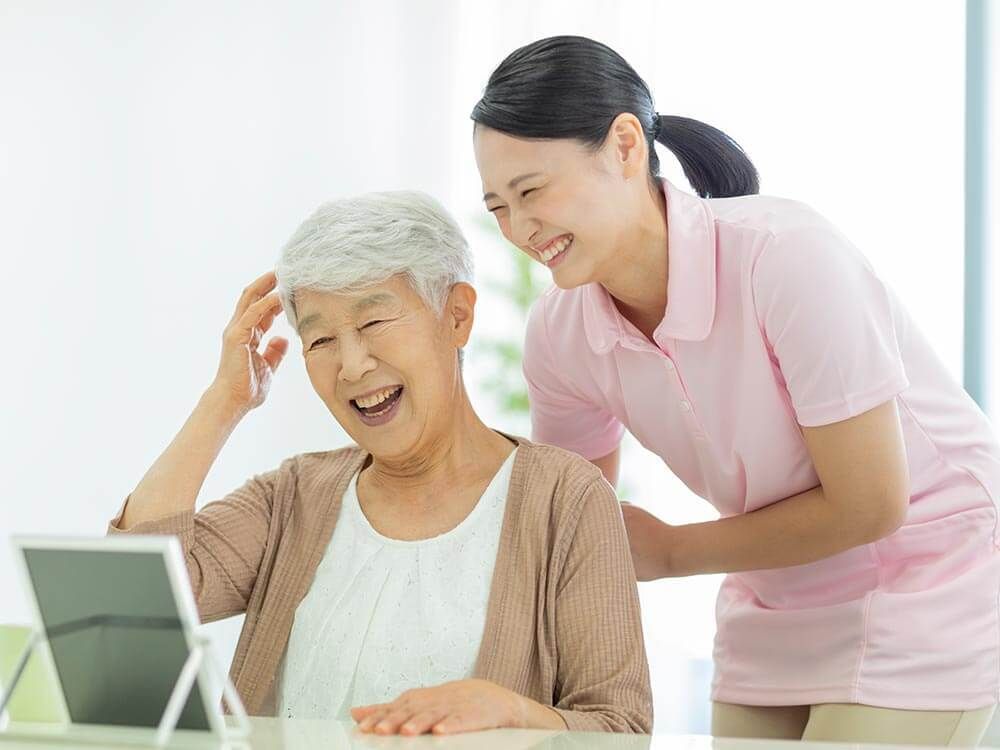 鏡を見ながら笑顔で話している高齢者とそこに寄り添介護スタッフの様子。明るい室内の写真
