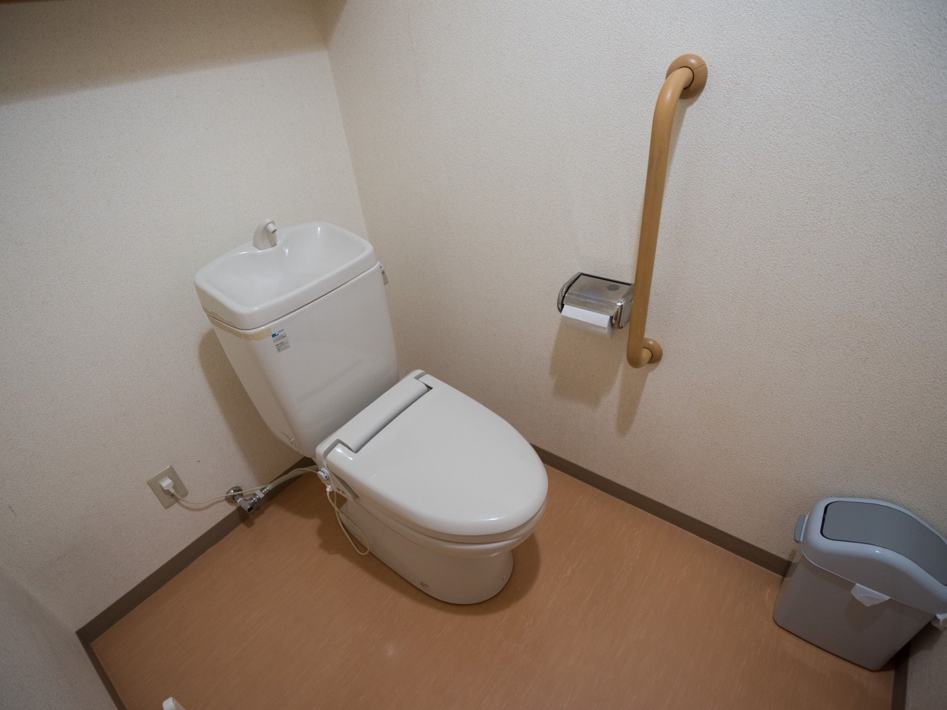 広々としたトイレである。洋式便座を完備。壁には縦型の手すりが備え付けられている。グレーのゴミ箱もある。