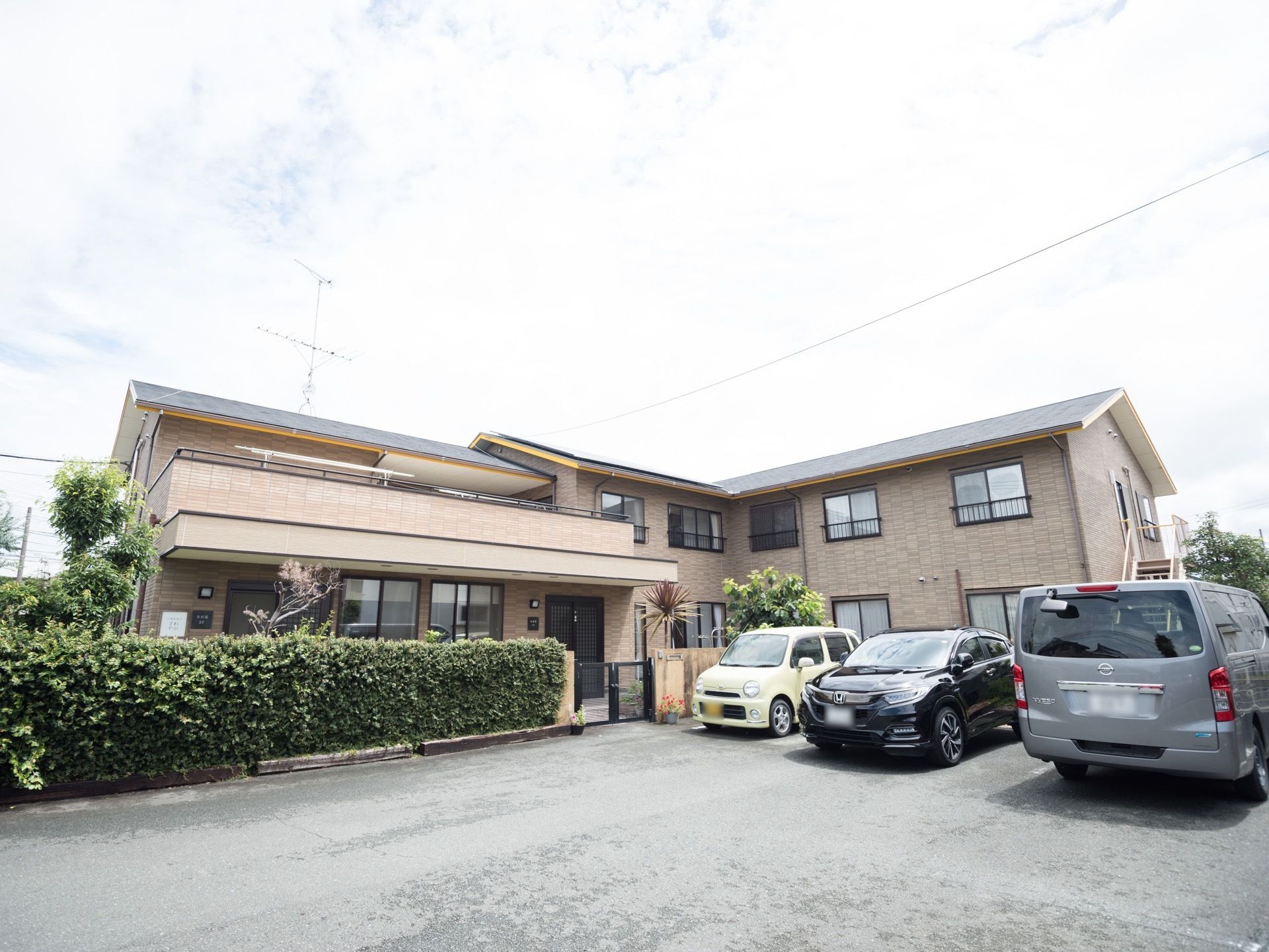 落ち着いた色合いの２階建て施設である。こまめに整備された、庭木が植えられている。敷地には車が複数台停められている。