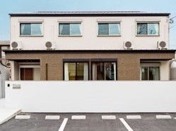 2階建ての建物の1階部分が茶色、2階部分がベージュ。建物の前には壁があり、駐車場もある。
