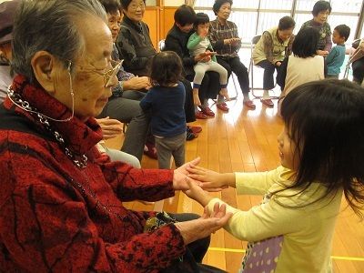 小さな女の子との手を取り、微笑む眼鏡をかけた高齢の女性。輪になって椅子に座る高齢者の元を、たくさんの子供たちが訪れている。