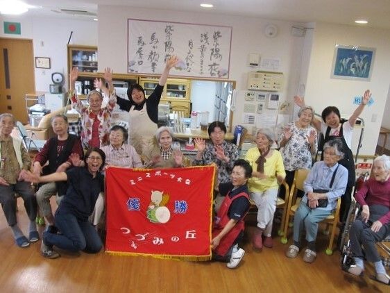 ホーム名が書かれた赤い旗を掲げ、満面の笑顔でスタッフと入居者たちが記念撮影に応じている様子。