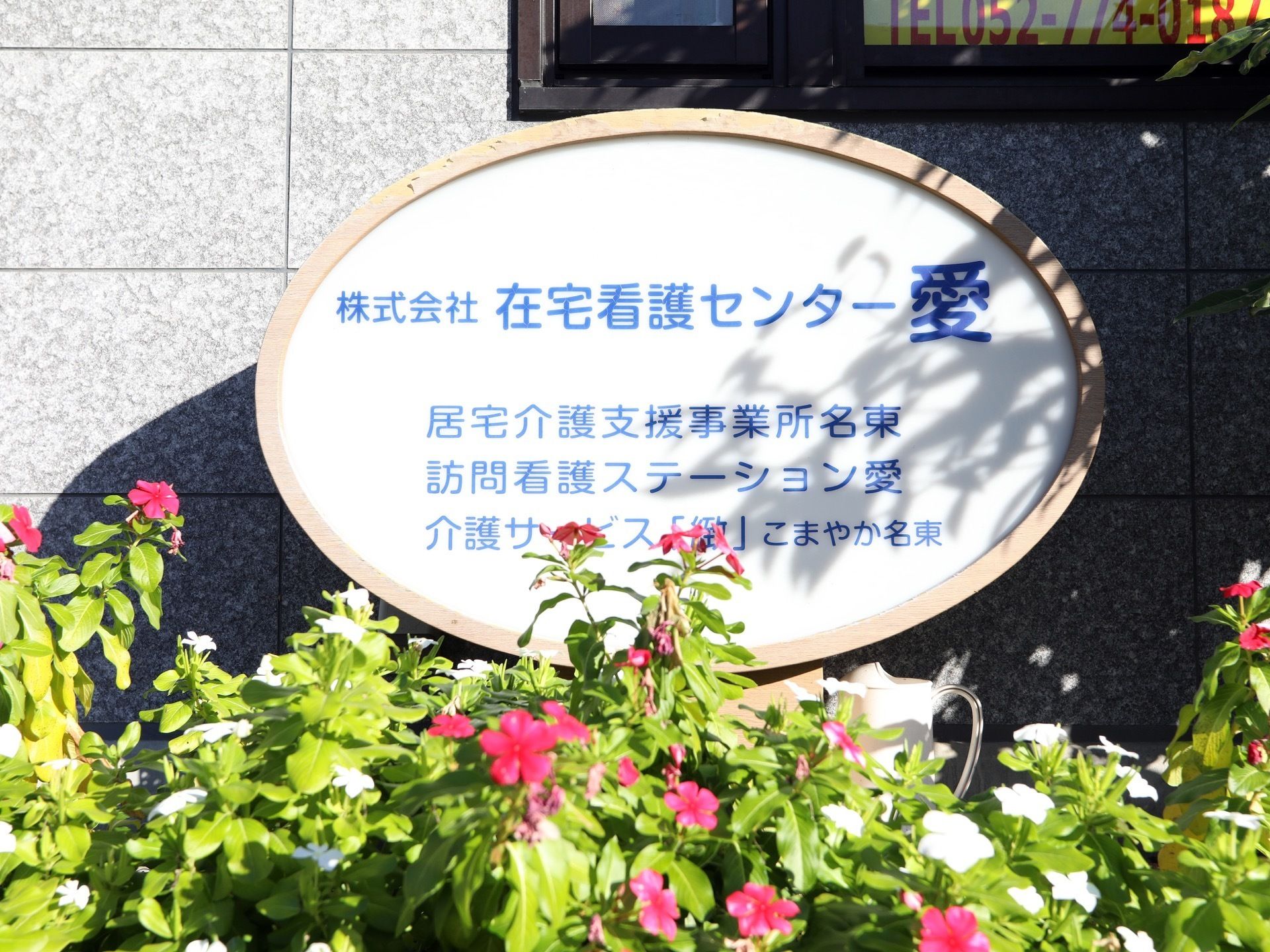 玄関前には、施設名の書かれた白くて丸い表札が設置されている。周りには、ピンク色の花がたくさん植えられている。