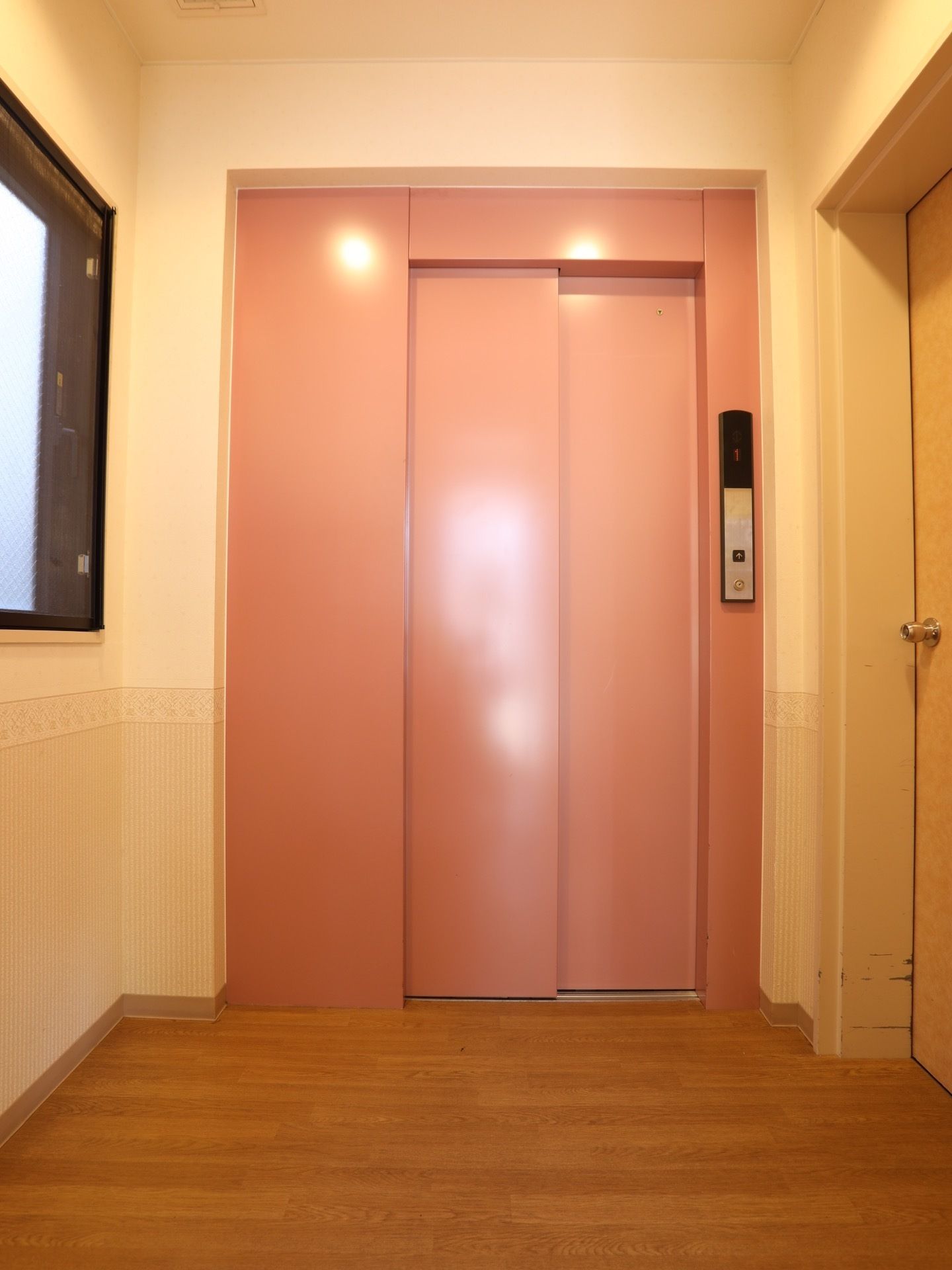 廊下は、全面フローリングの床となっている。奥には、ピンク色の扉がついたエレベーターが完備されている。