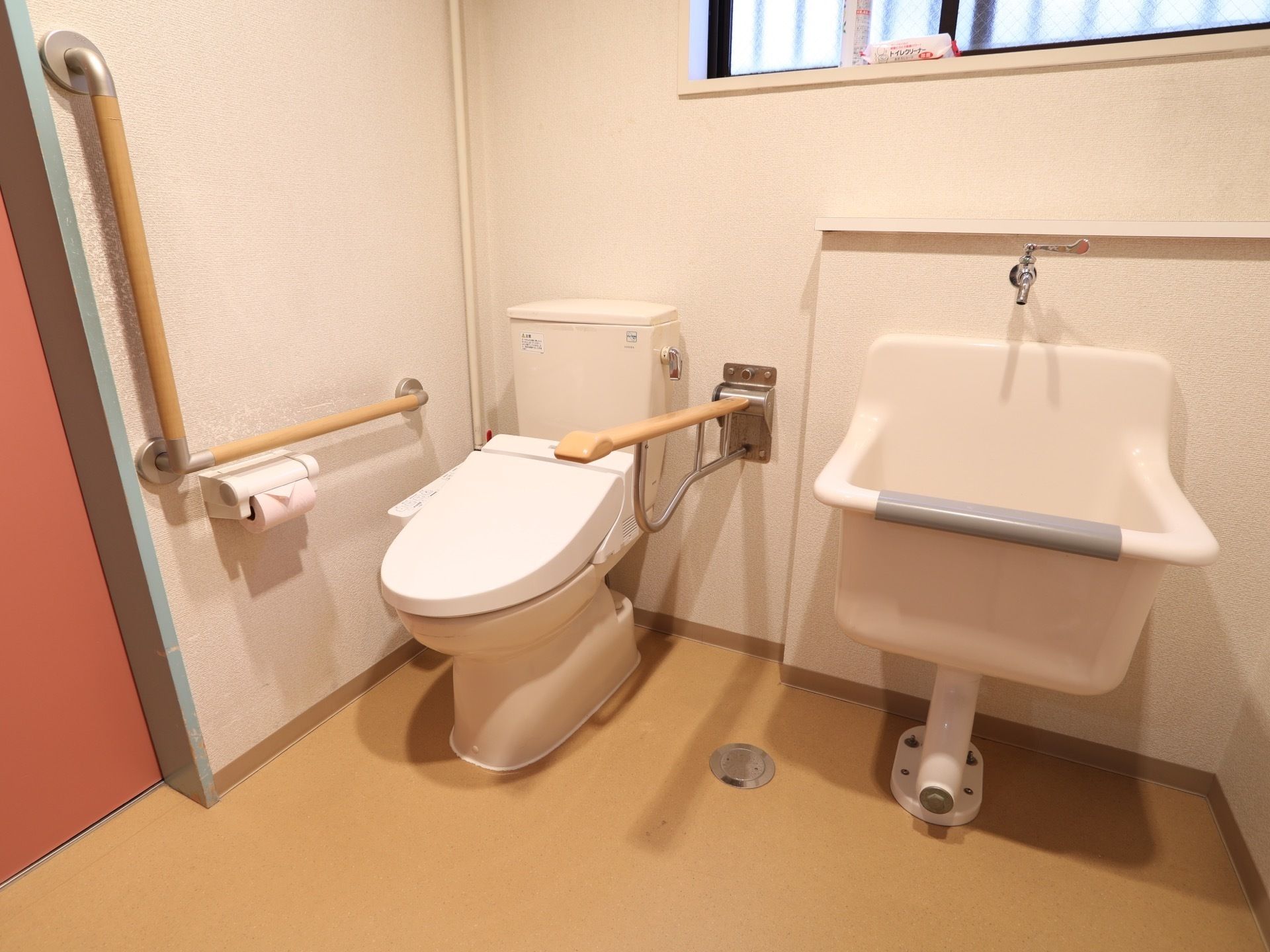 施設には、ピンク色の扉が取りつけられたトイレがある。壁には茶色い手すりもあり、手洗い場も設置されている。