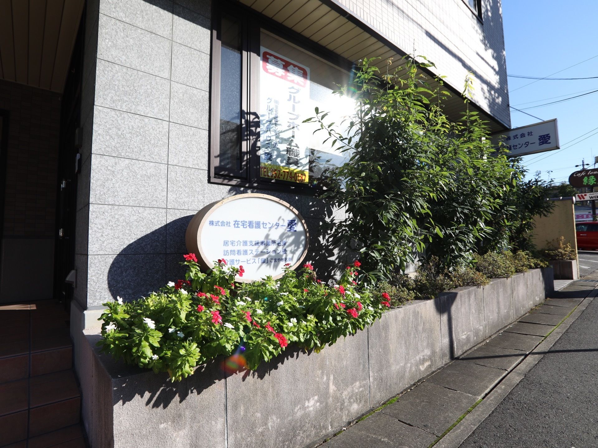 施設の外壁はグレーで、玄関前にはたくさんの植物が植えられている。その左端には、丸い形の表札が設置されている。