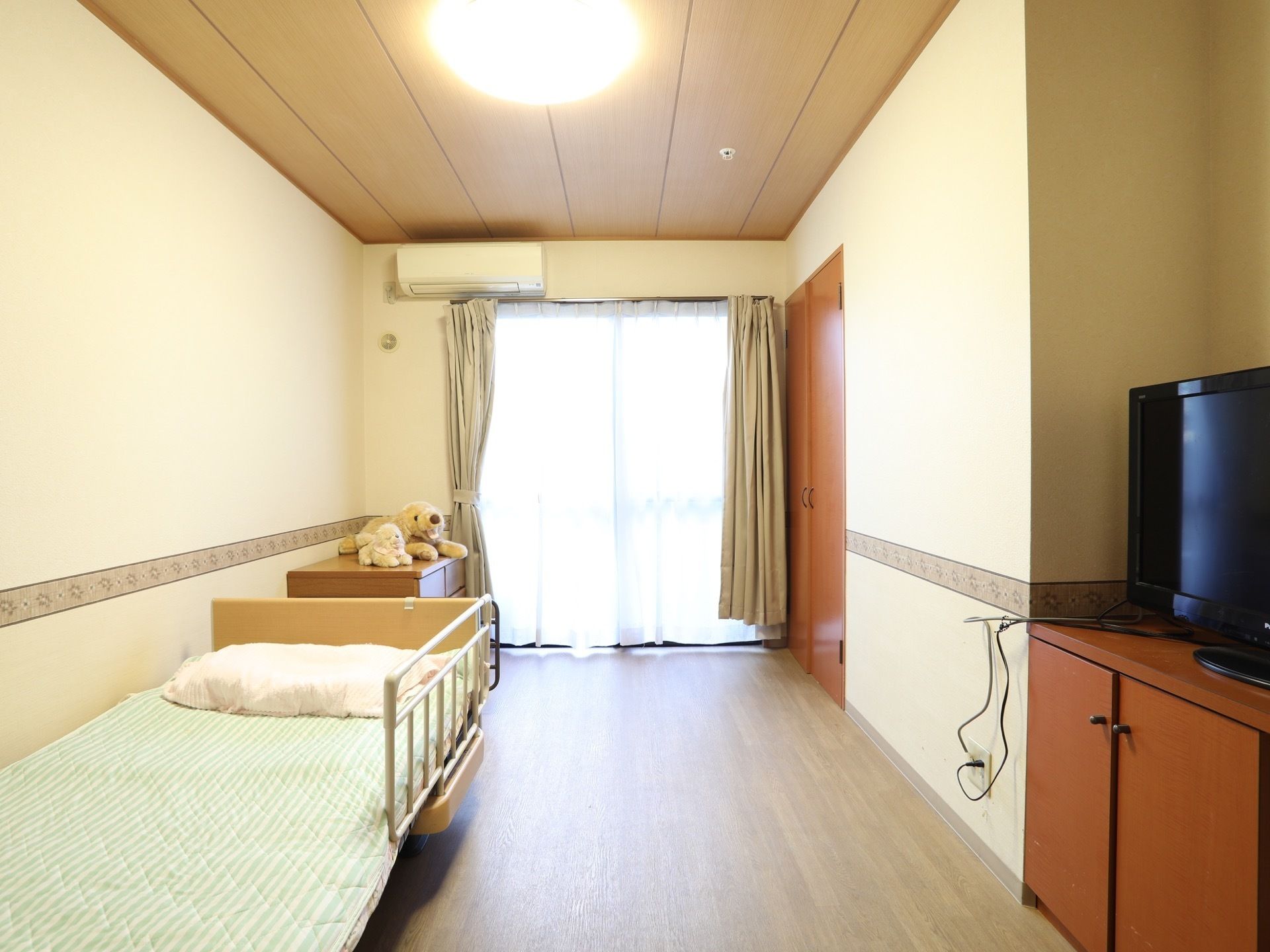 居室は奥行きがあり、左側にはタンスとシングルベッドが置かれている。右側の収納棚の上には、テレビが設置されている。