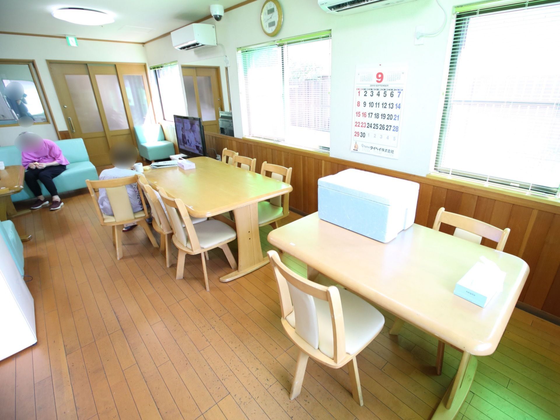ダイニングテーブルが２つといくつもの椅子が置かれている食堂。天井近くの壁にはエアコンが２つ備えられている。