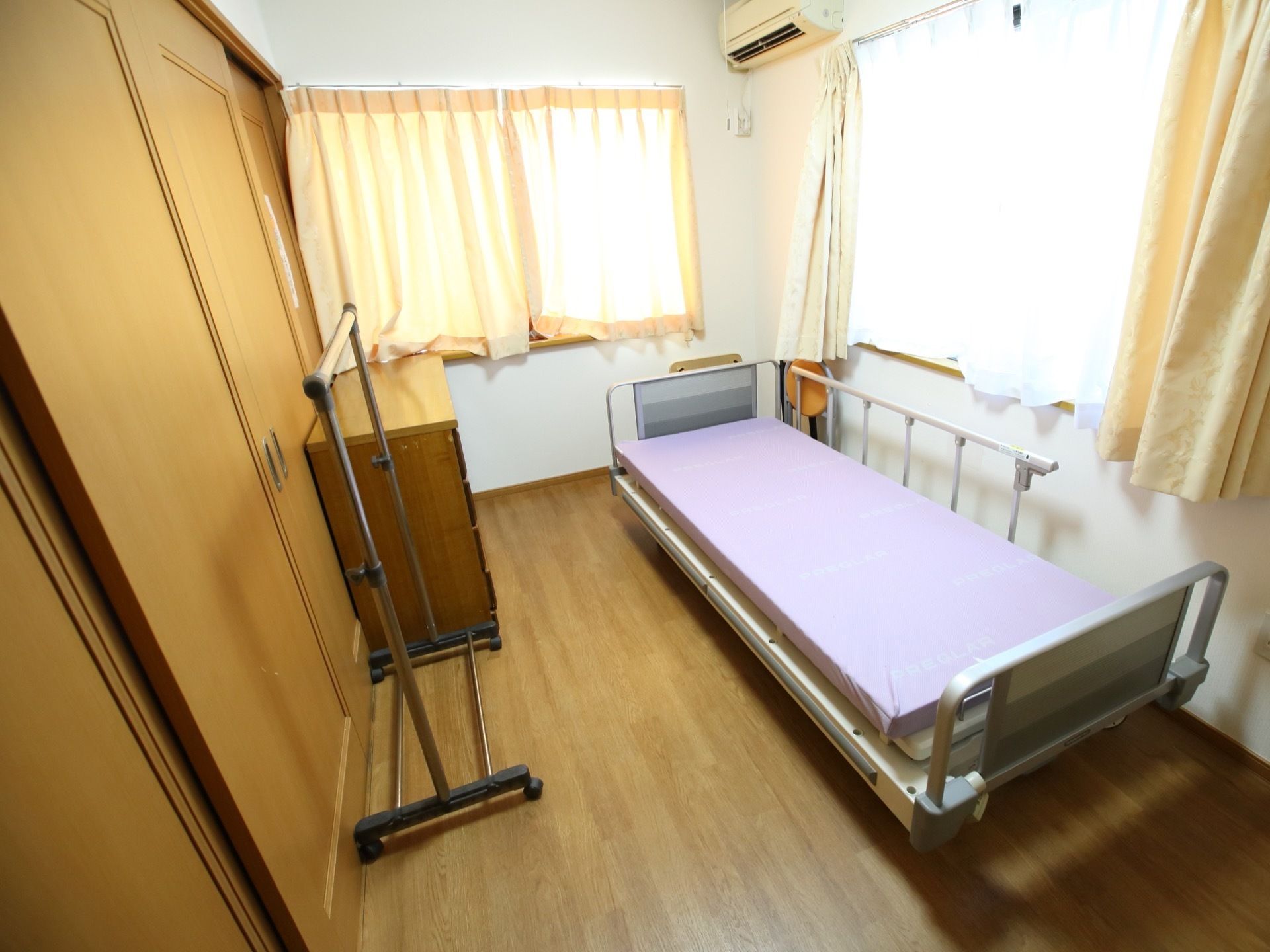 ２つの窓がある明るい居室。窓と窓の間の壁に、エアコンが設置されており、介護用ベッドや収納などが備えられている。