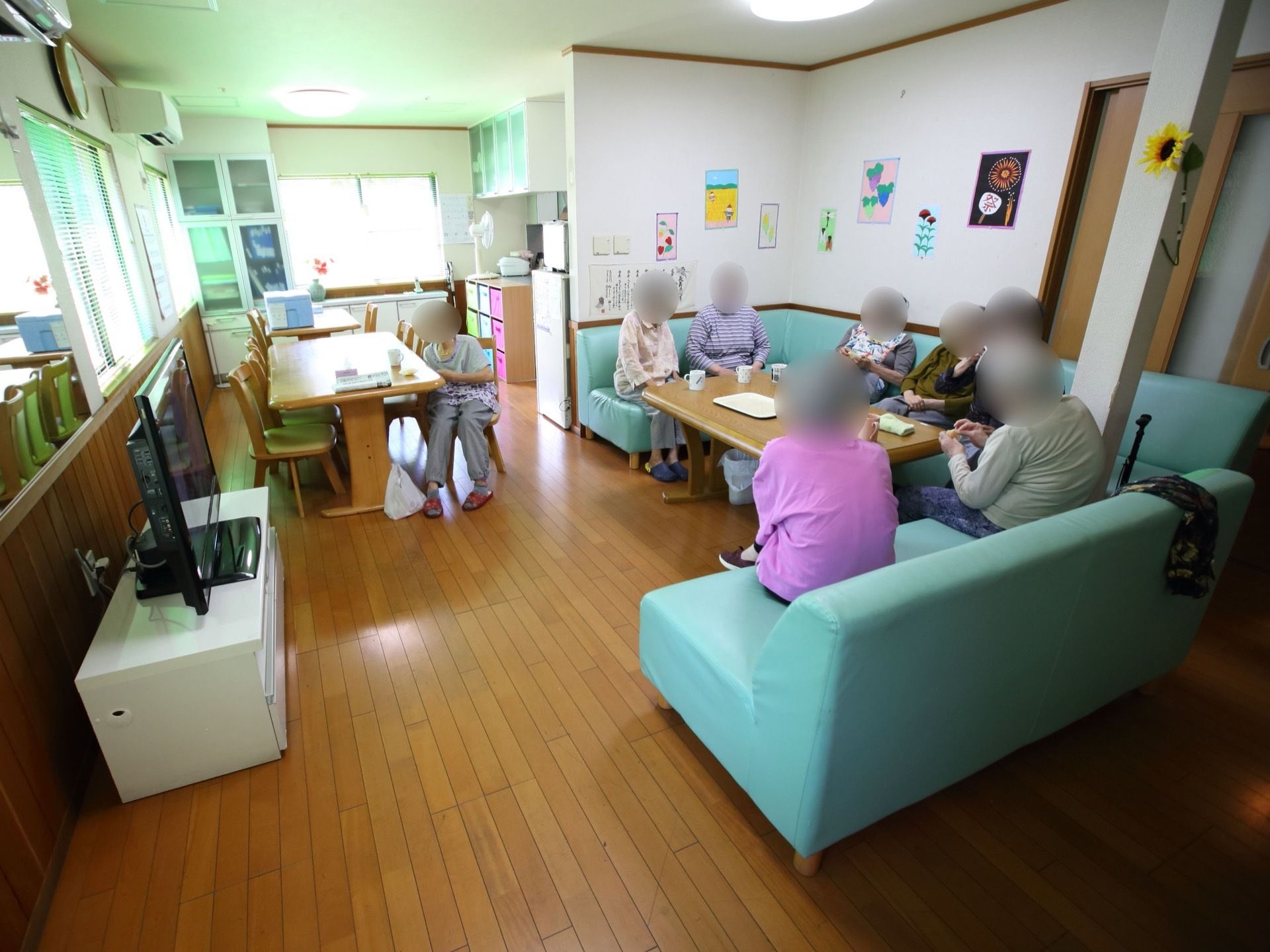 テレビとソファ、テーブルなどが設置された喫茶店のような内装のリビング。ソファに座ってお茶を飲んでいる人々がいるのが見える。