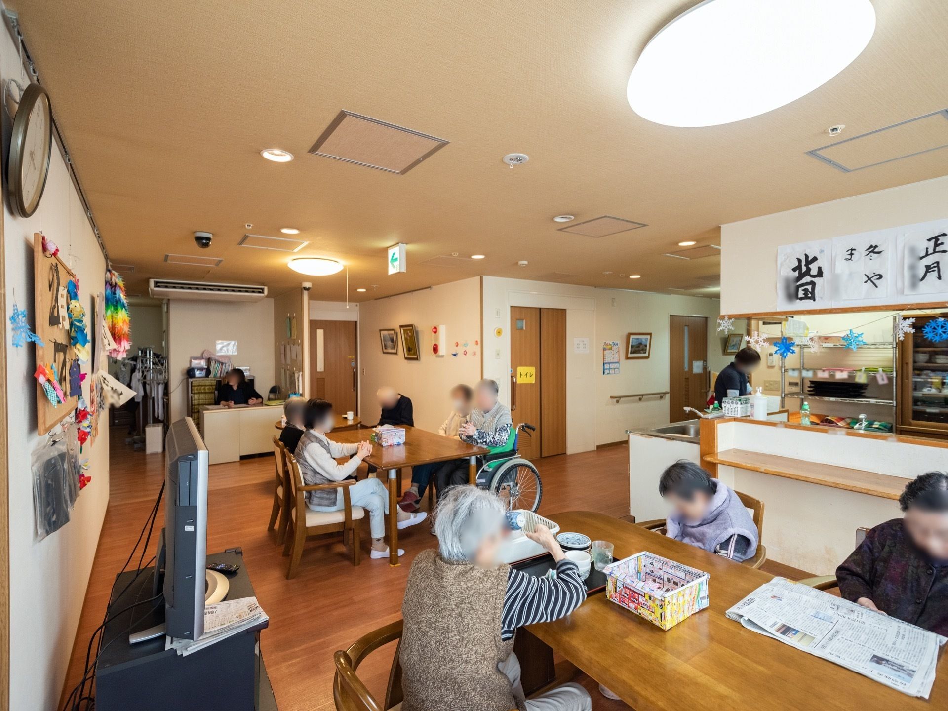 右奥にはキッチンがあり、スタッフが作業をしている。ダイニングにあるテーブルセットでたくさんの入居者様がくつろいでいる。