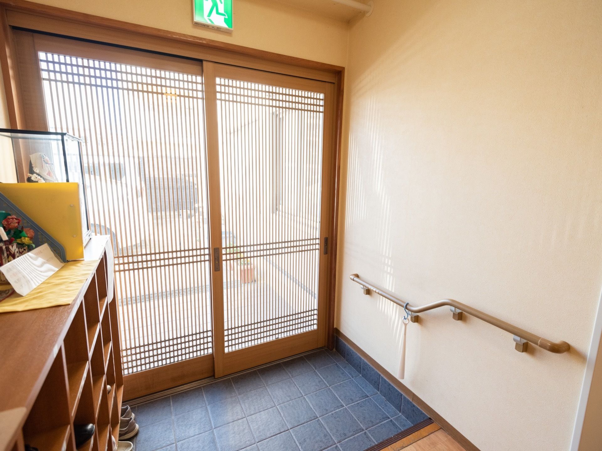 細い木製の格子がはまった入口扉が正面にあり、その上に非常口の電気がついている。左手前に下駄箱がある。